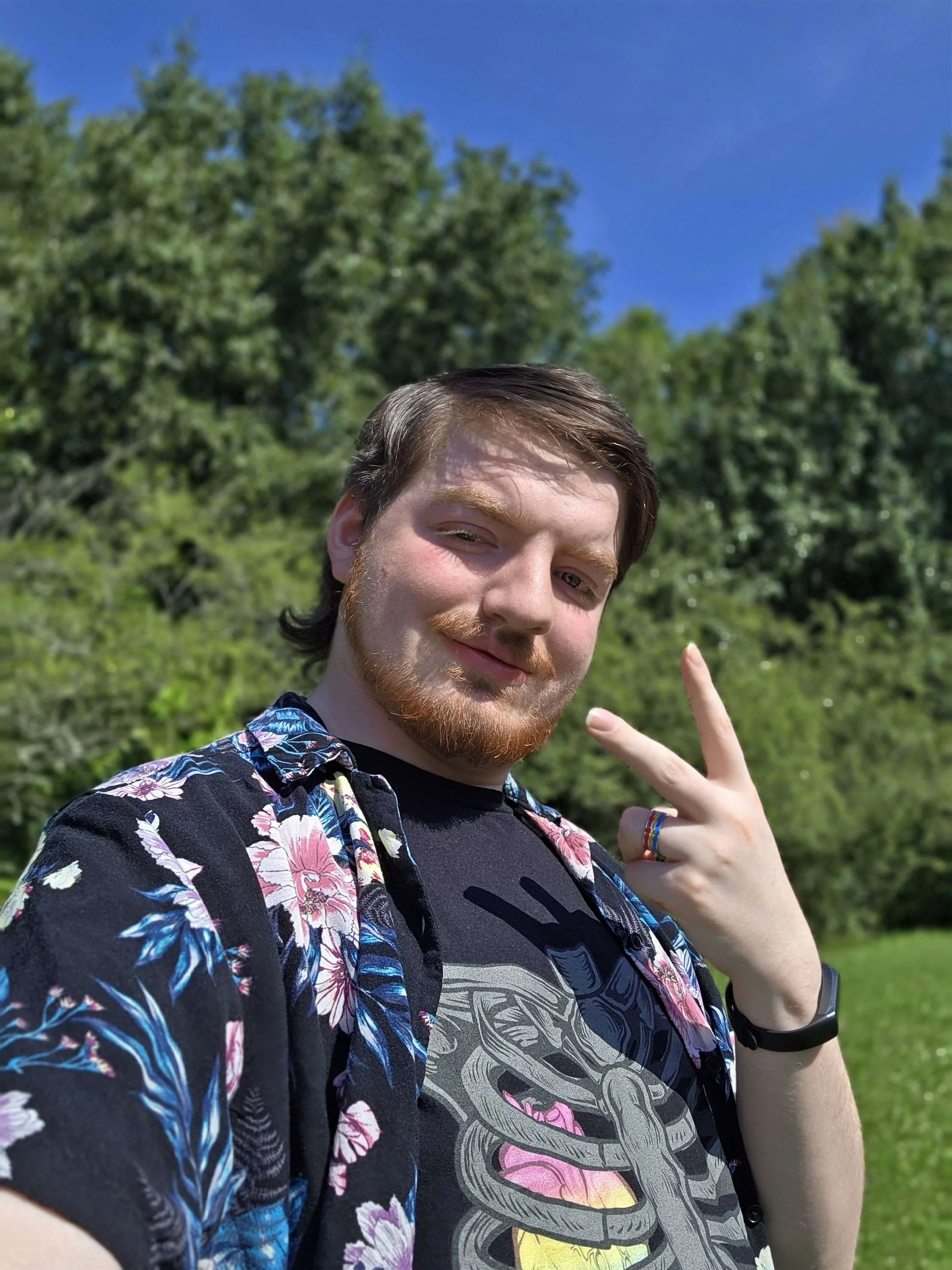 A man with brown hair and a beard outdoors, smiling and flashing a peace sign, wearing a black ribcage t-shirt with a hawaiian shirt on top of it, a pink-yellow-blue banded ring, and a black watch, with green trees and a blue sky in the background.