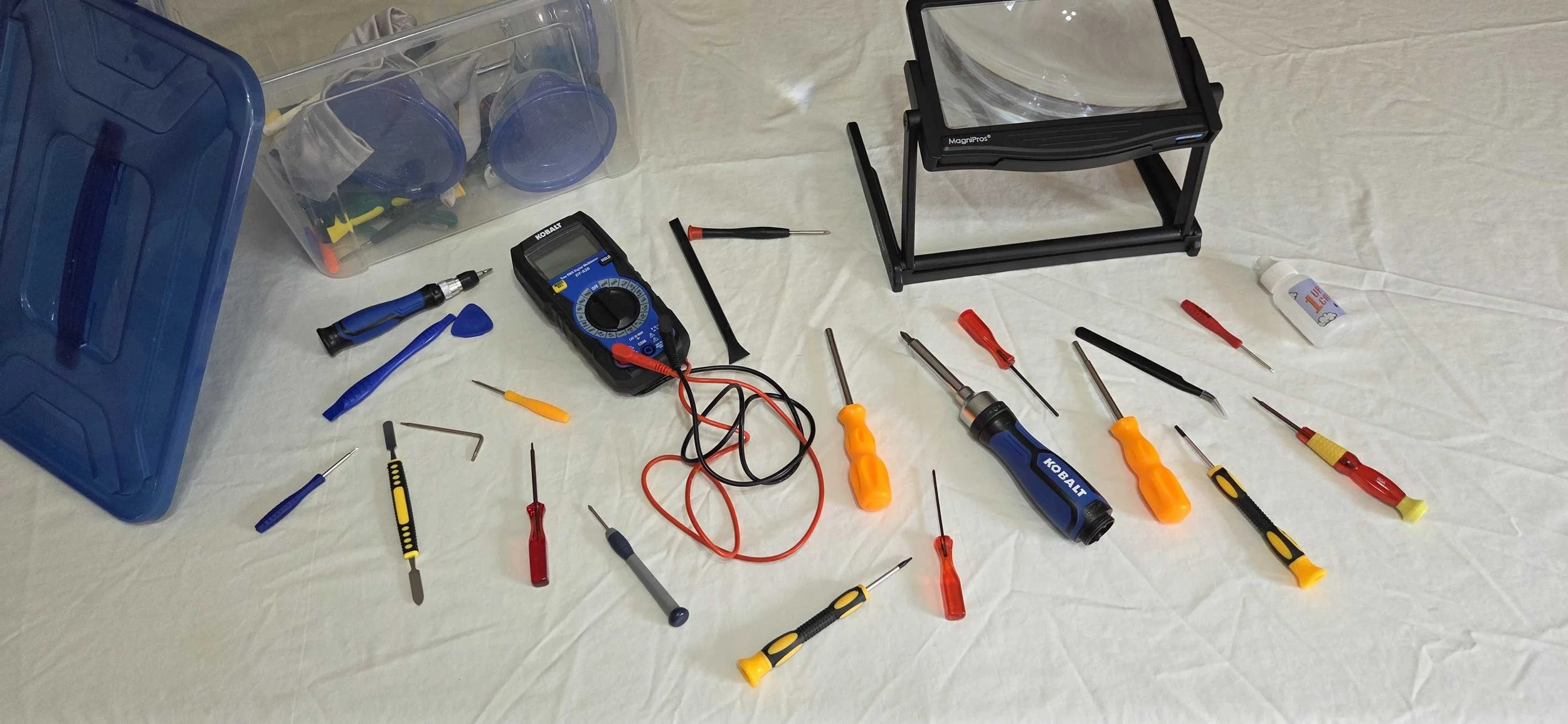 Assorted electronic repair tools, including screwdrivers, a multimeter, tweezers, and small containers for parts, arranged on a white cloth with a blue storage box and a black magnifying glass stand.