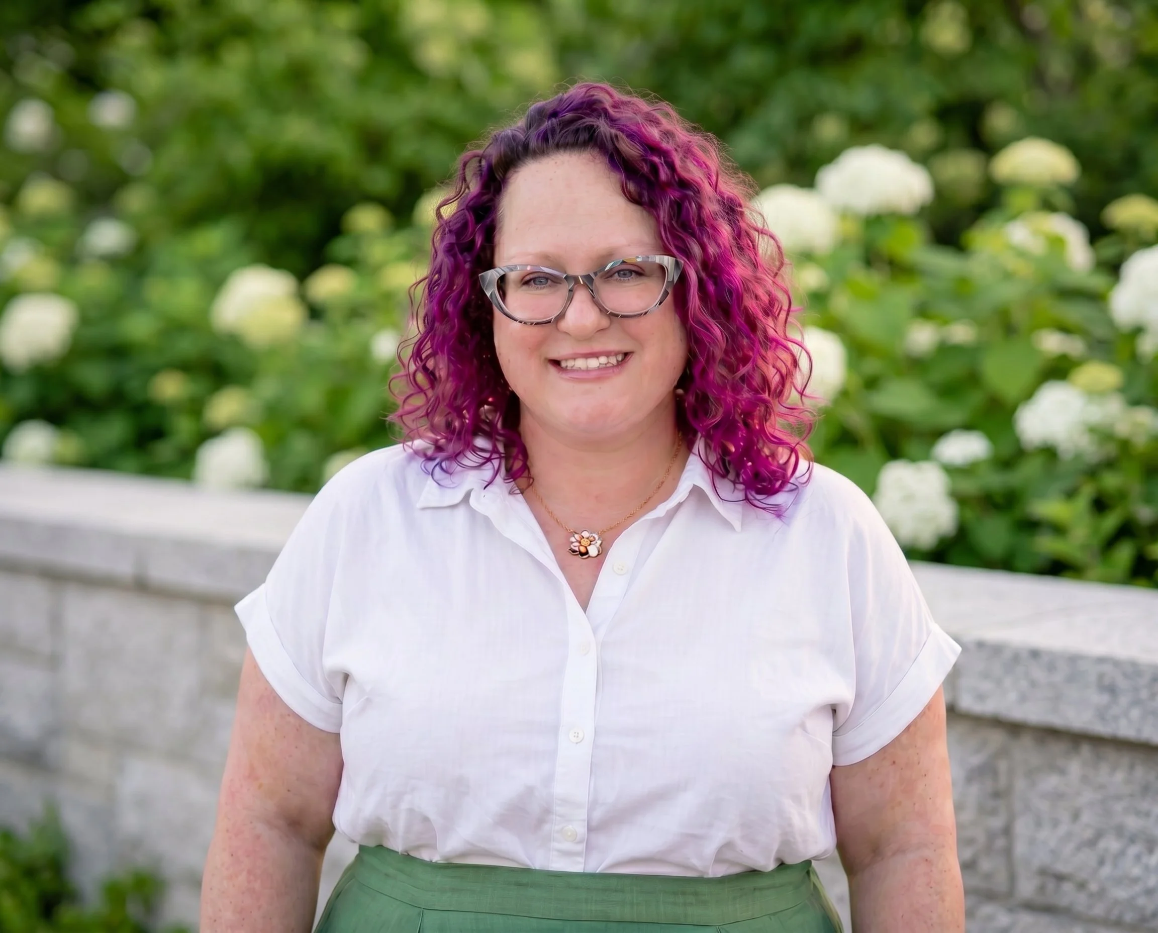 A picture of Kate with curly purple and pink hair, glasses, wearing a white blouse and green skirt, standing outdoors with a blurred green and white background.