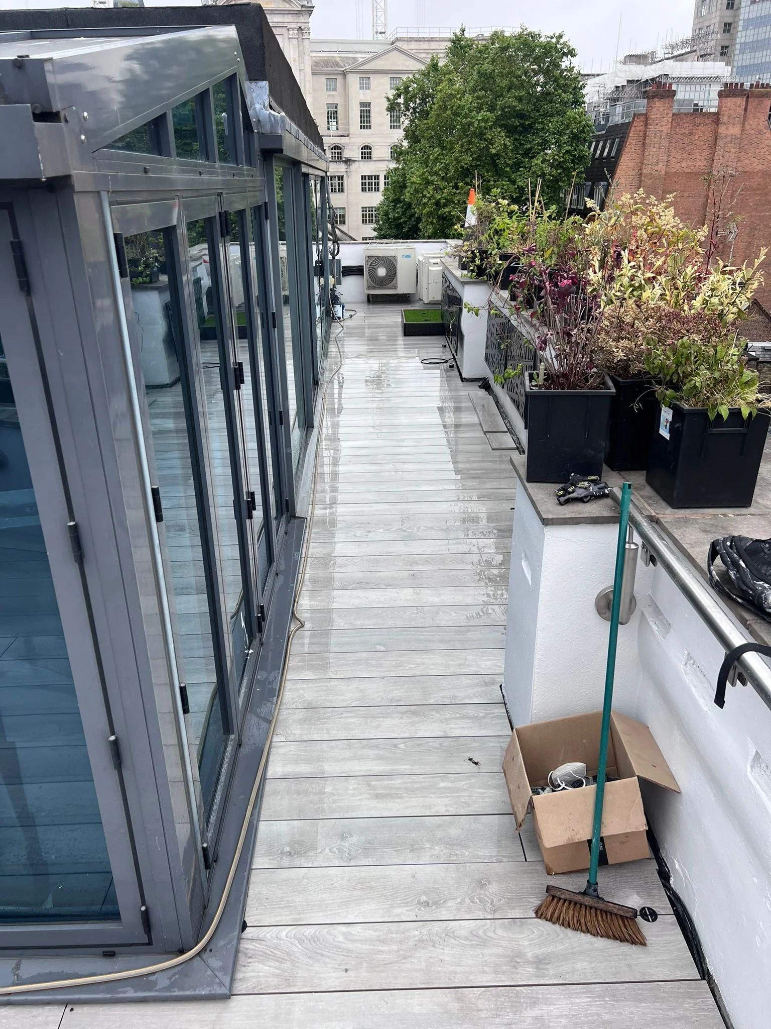 A rooftop balcony with a glass-enclosed area on the left, outdoor heating and cooling units at the end, potted plants with purple and white flowers along the railing, a broom and dustpan, an open cardboard box with miscellaneous items, and a wet wood