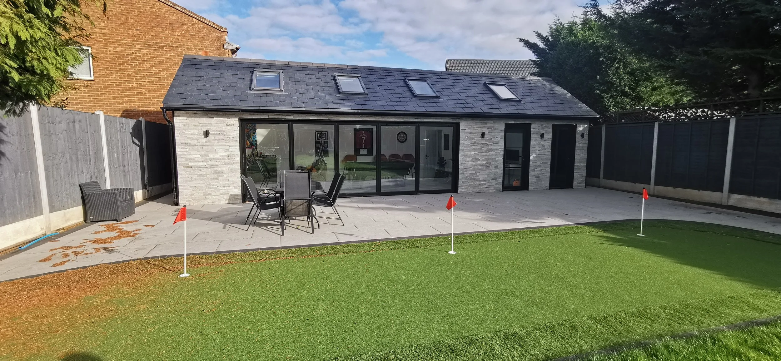 Backyard patio with outdoor table and chairs, artificial turf putting green, and a modern house with large glass doors and skylights on the roof.
