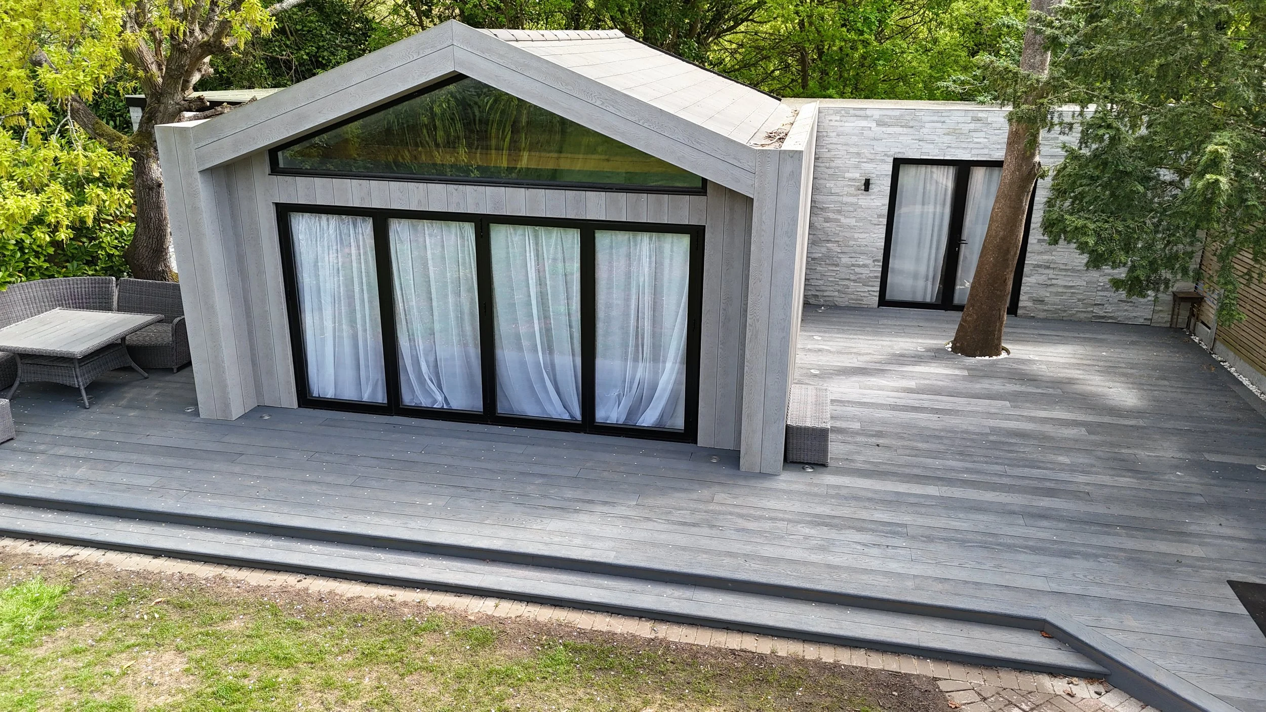 Modern house with large glass sliding doors and windows, situated on a wooden deck surrounded by trees.