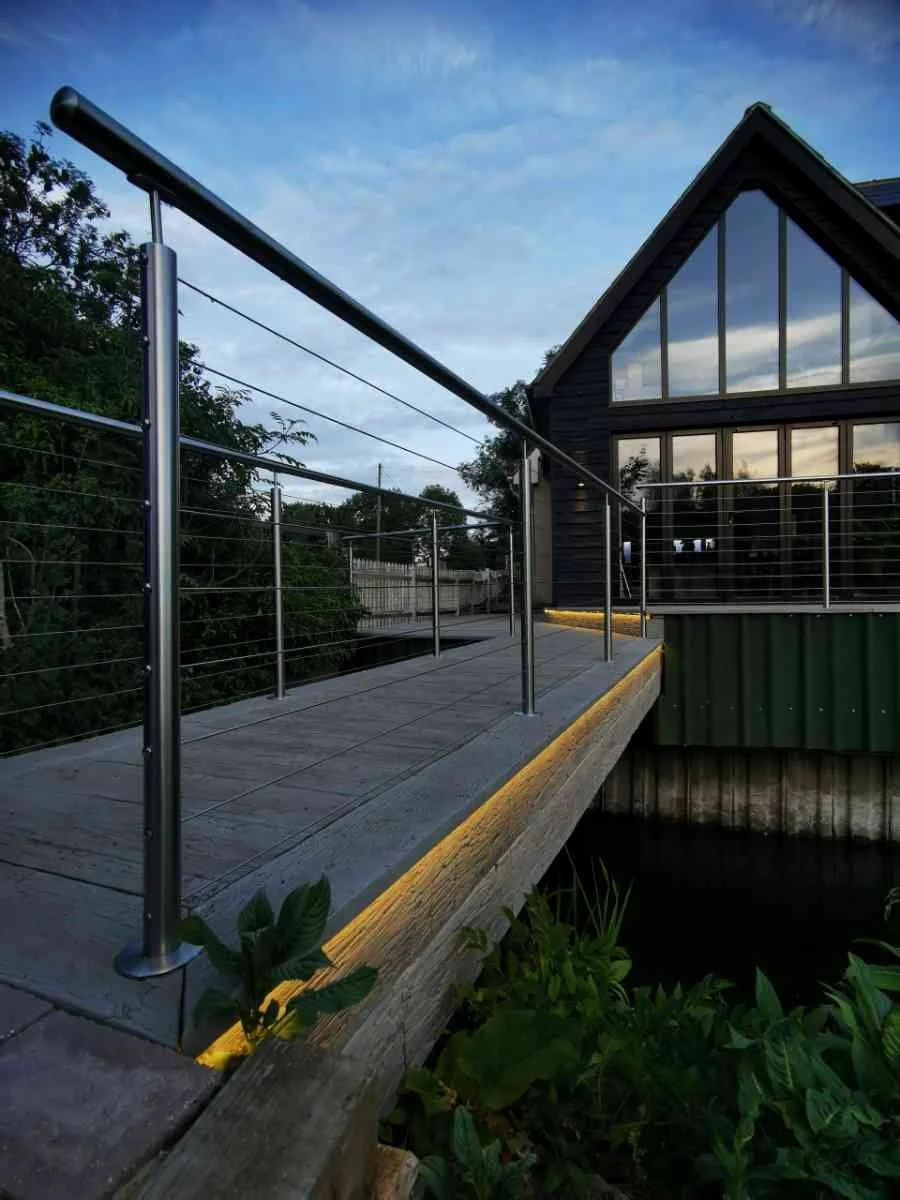 A wooden deck with metal cable railings in front of a modern house with large glass windows, surrounded by greenery, during dusk with a partly cloudy sky.