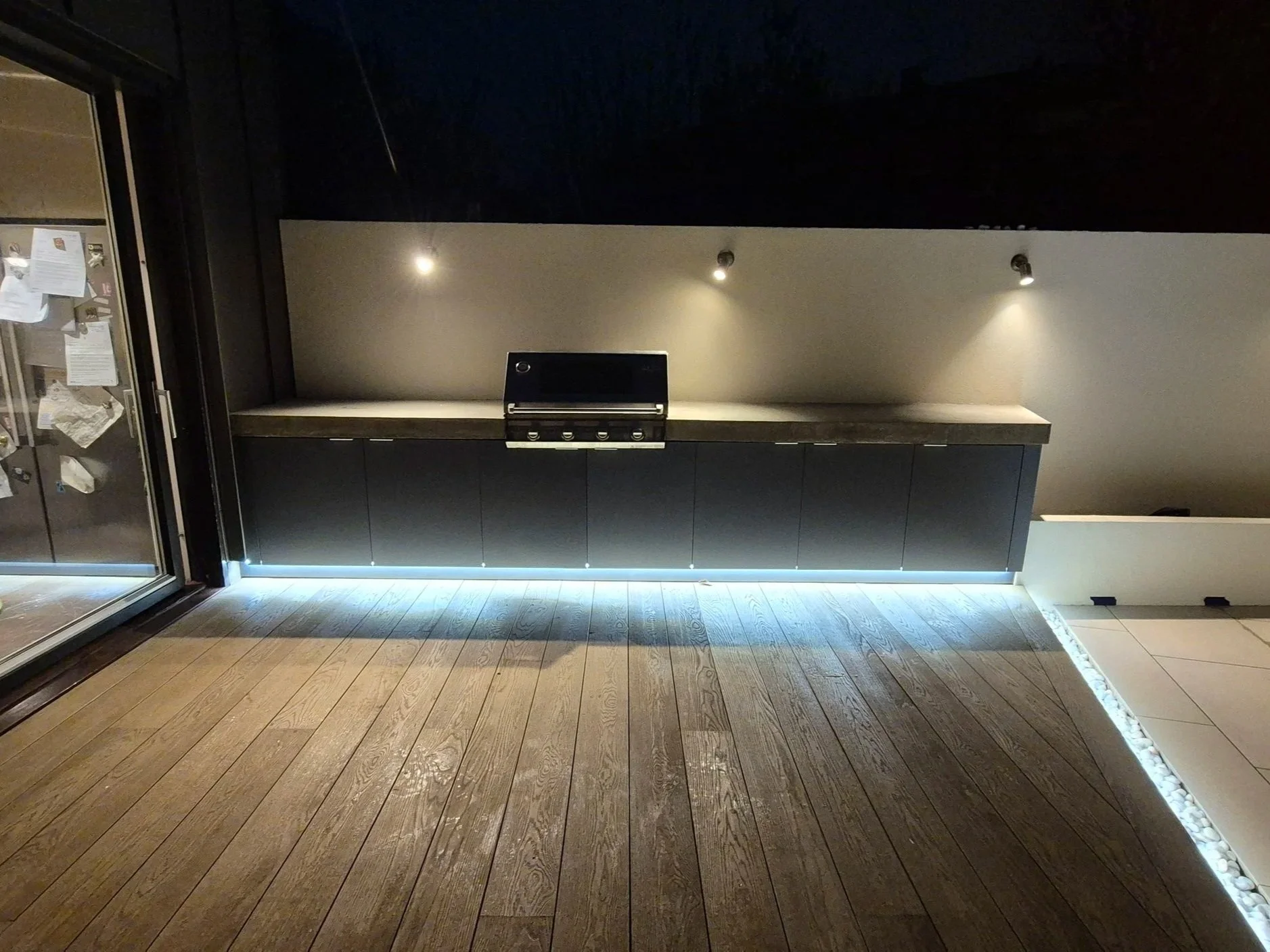 Backyard with wooden deck, modern black cabinet beneath a beige wall with three ceiling spotlights, and a strip of white stones along the edge of the deck.