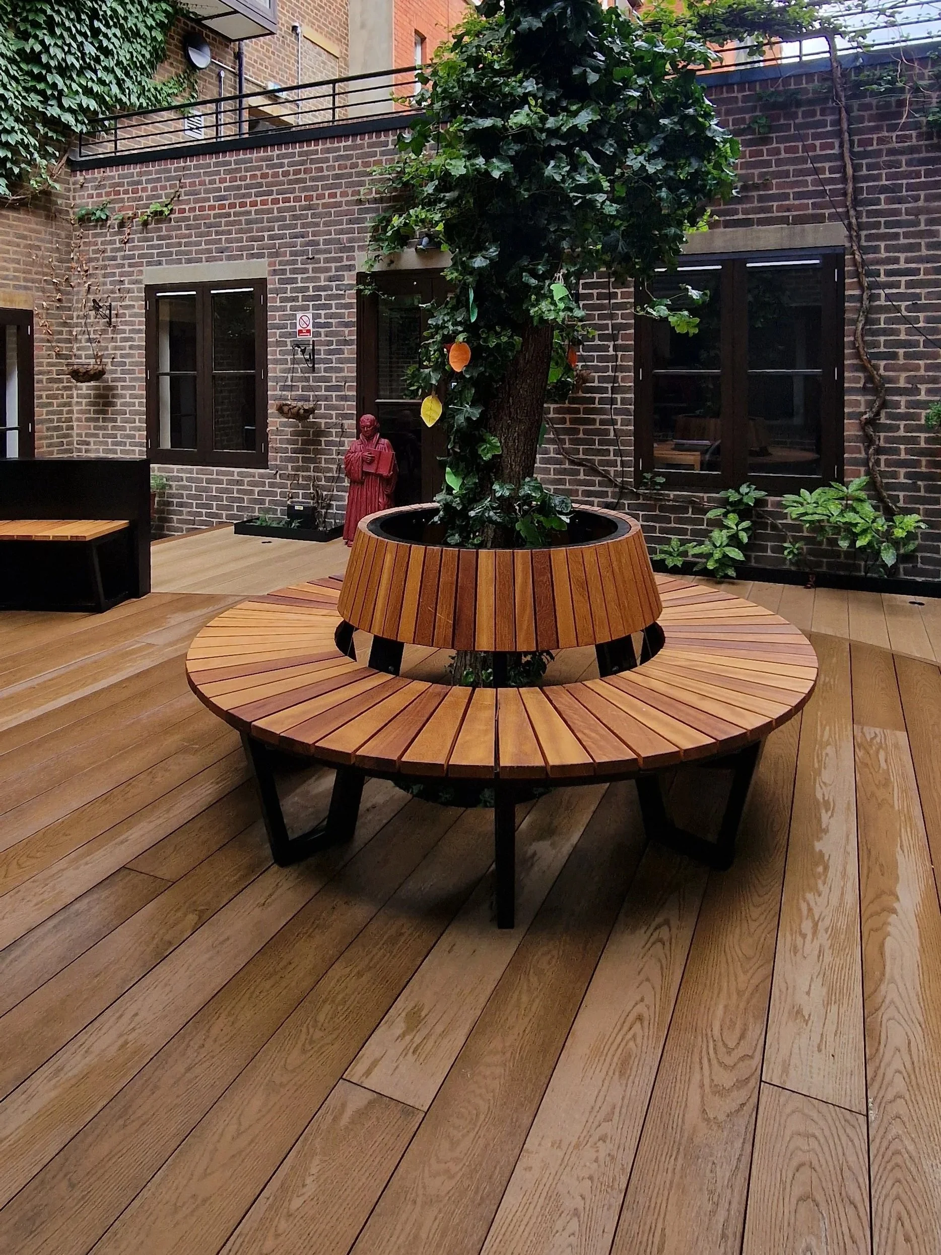 A brick-walled courtyard with a wooden circular bench surrounding a tree. The tree has green leaves and some colorful paper decorations hanging from it. The floor is made of wooden planks and there are two windows on the brick wall.