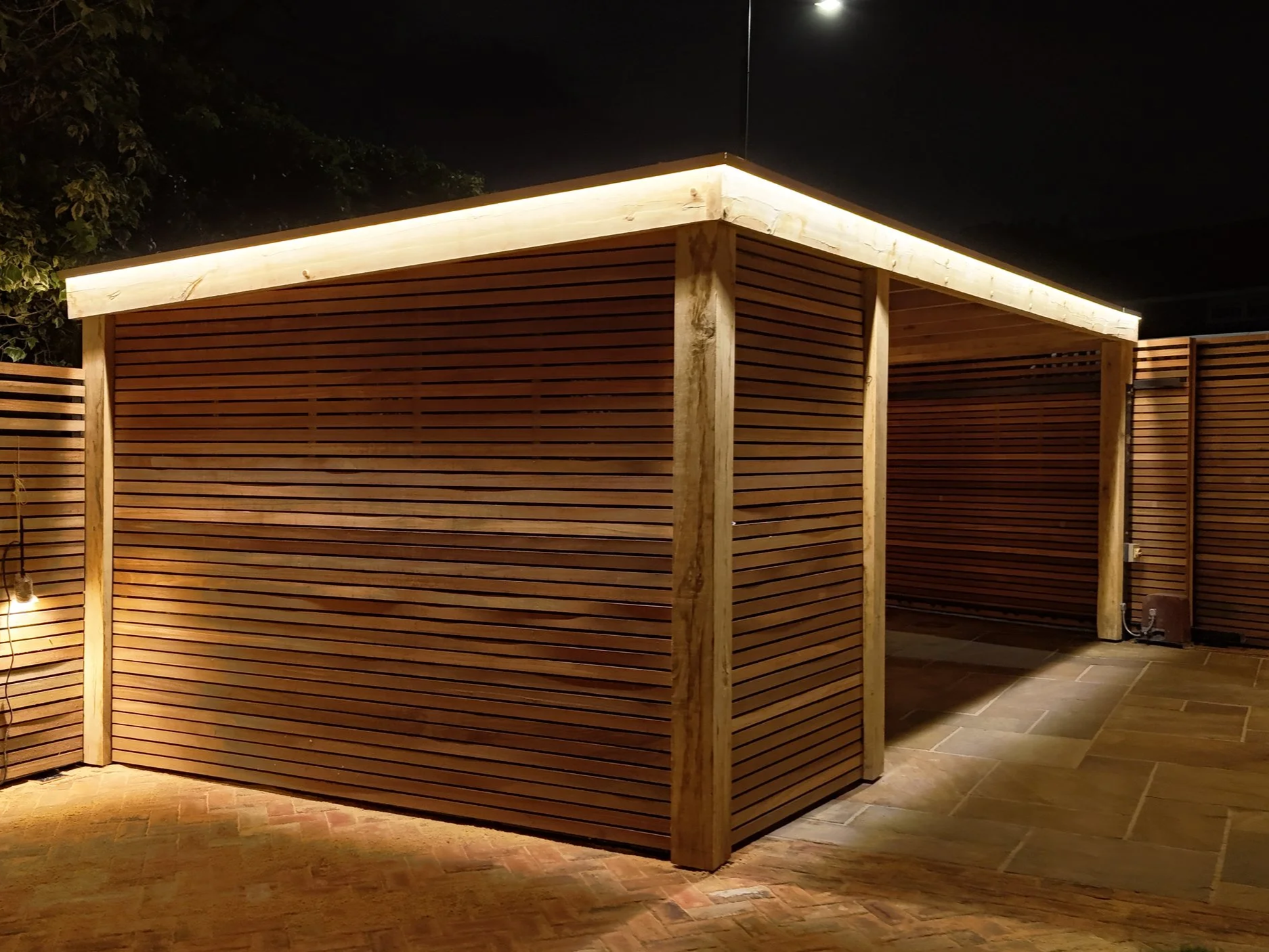 A wooden structure with slatted walls and a flat roof, illuminated with warm lighting at night, in an outdoor space.