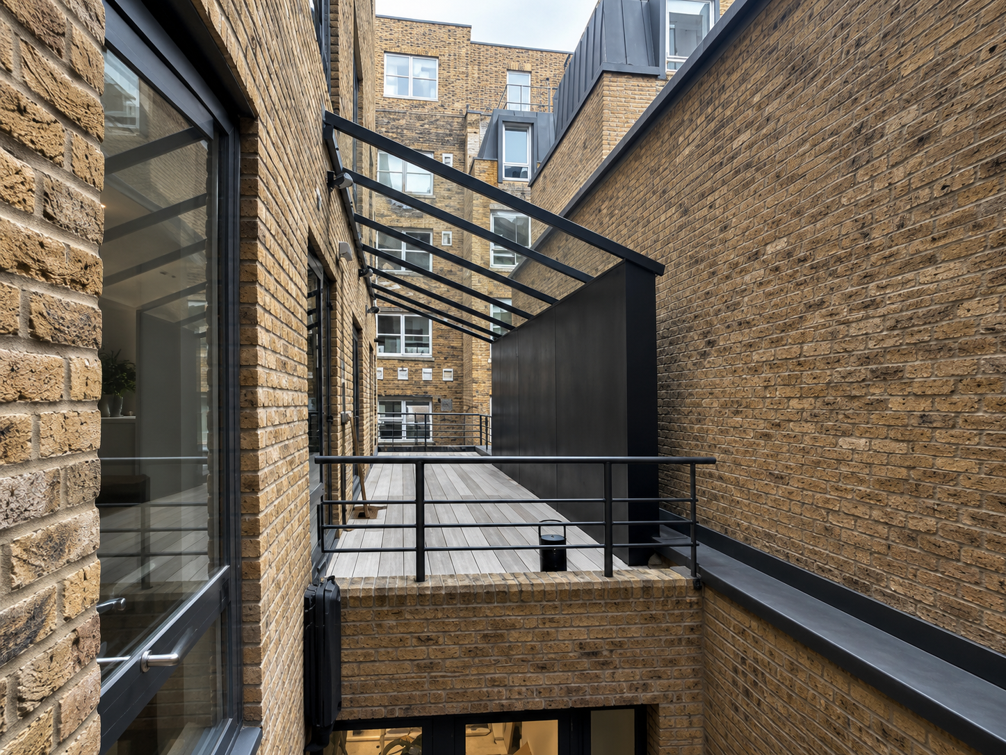 A narrow outdoor balcony with a wooden floor, enclosed by brick walls and black metal railings, connected to a brick building with large windows and glass doors.