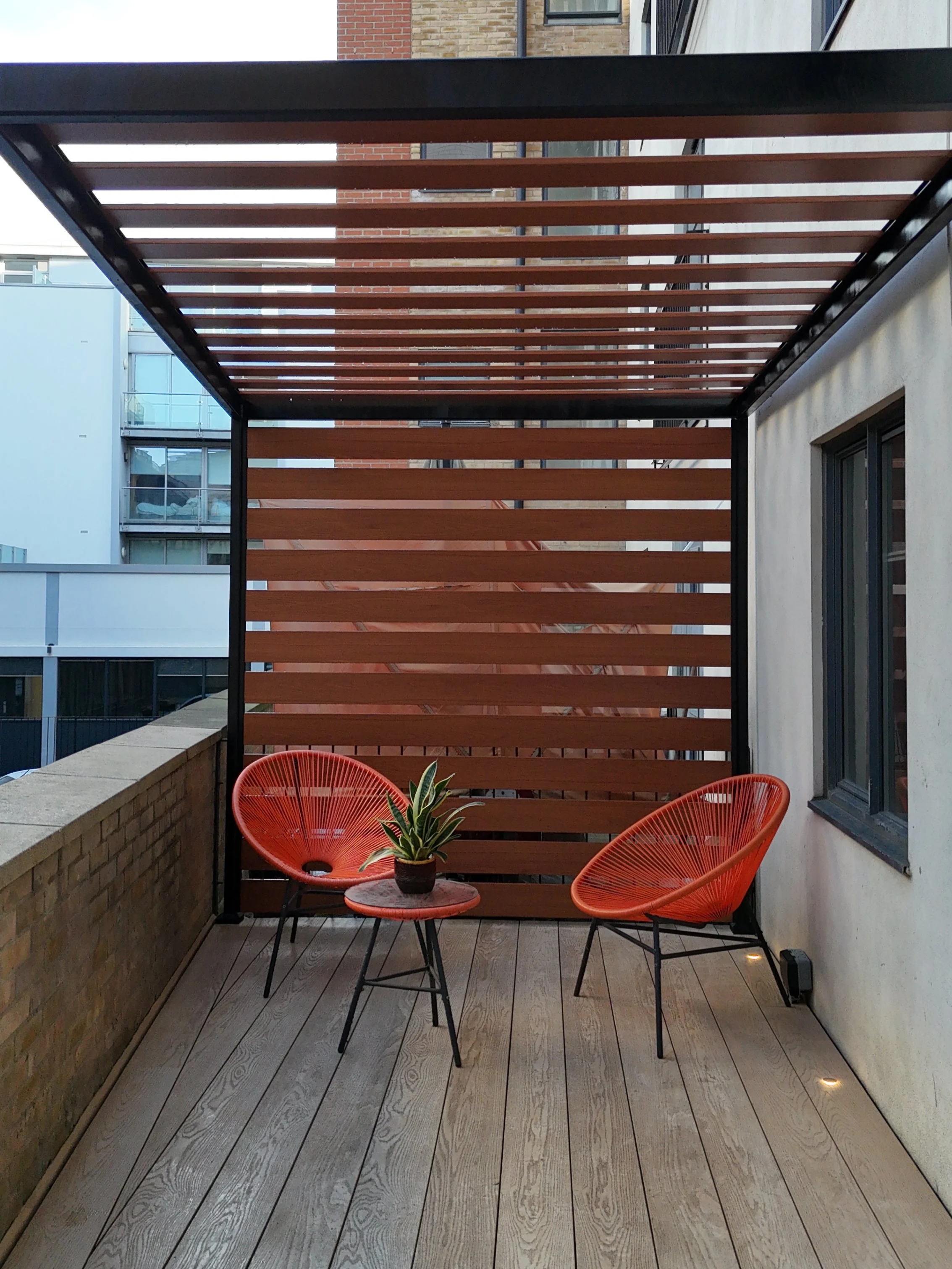 A small balcony decorated with two orange chairs, a small table, and a potted plant. The balcony has wooden flooring and a privacy wall made of horizontal wooden slats.