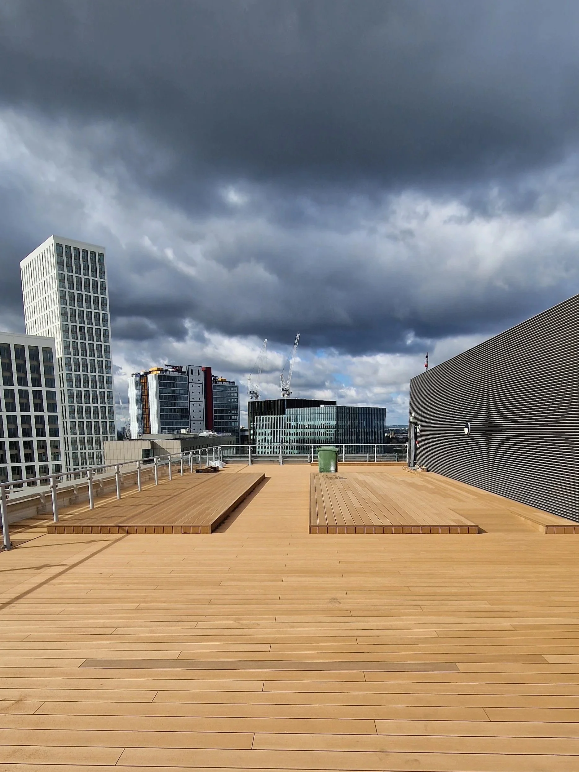 A rooftop terrace with wooden flooring and benches, surrounded by modern high-rise buildings under a cloudy sky.
