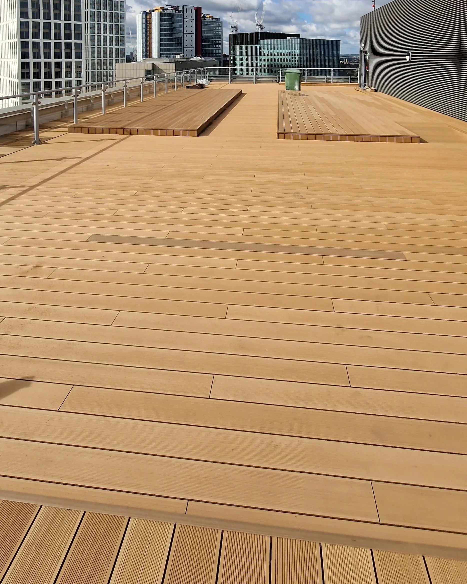 Rooftop terrace with wooden decking, surrounded by metal railings, and modern high-rise buildings in the background under a partly cloudy sky.