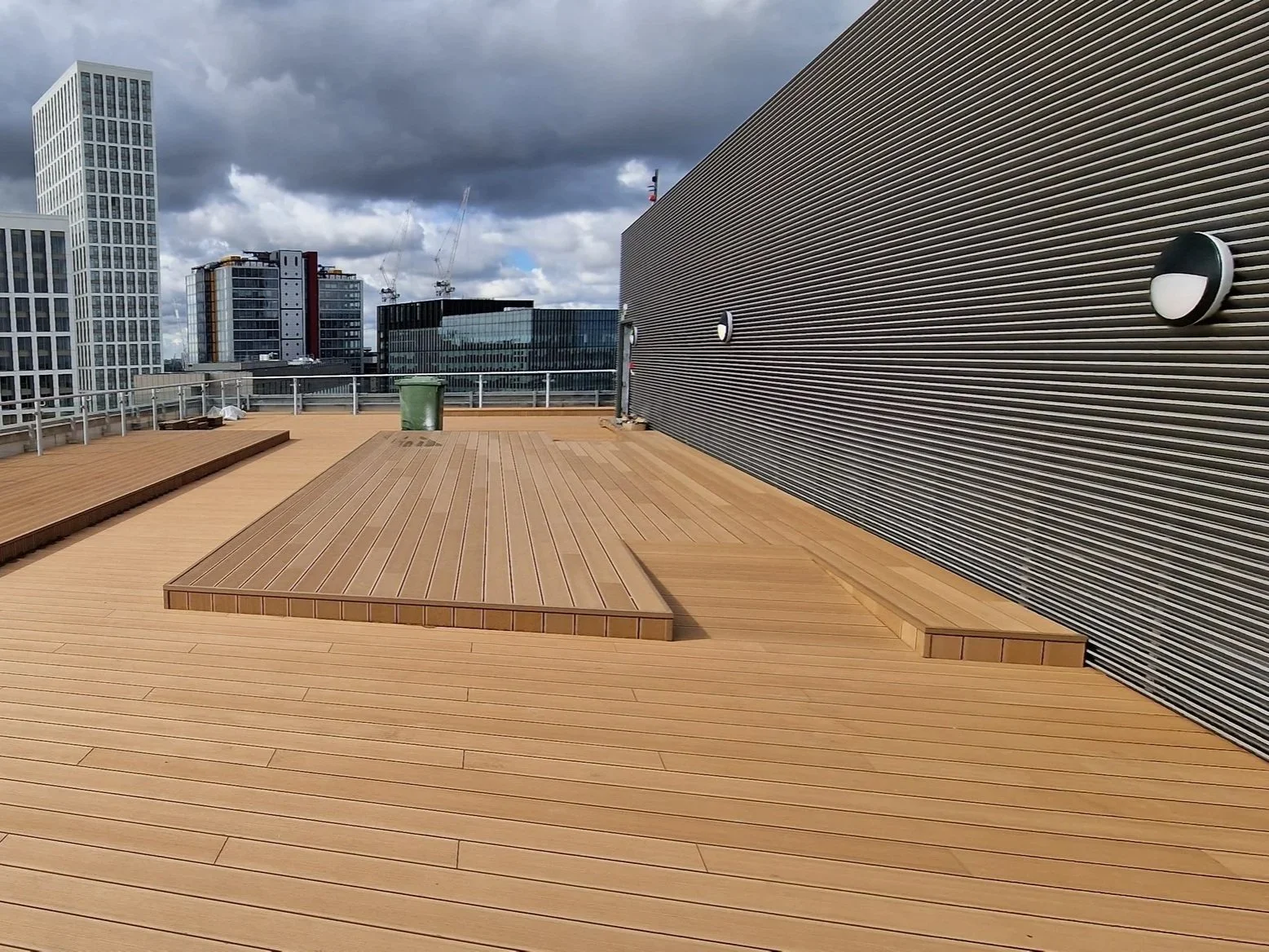 Empty rooftop deck with wooden flooring, a green trash bin, and a cloudy sky with high-rise buildings in the background.