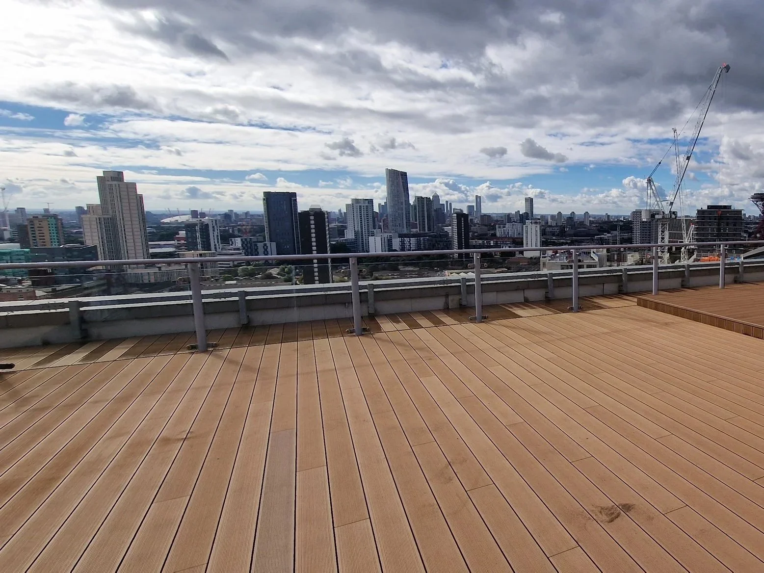 Rooftop view of a city skyline with high-rise buildings, a cloudy sky, and a crane in the background. The foreground features a wooden deck with metal railings.