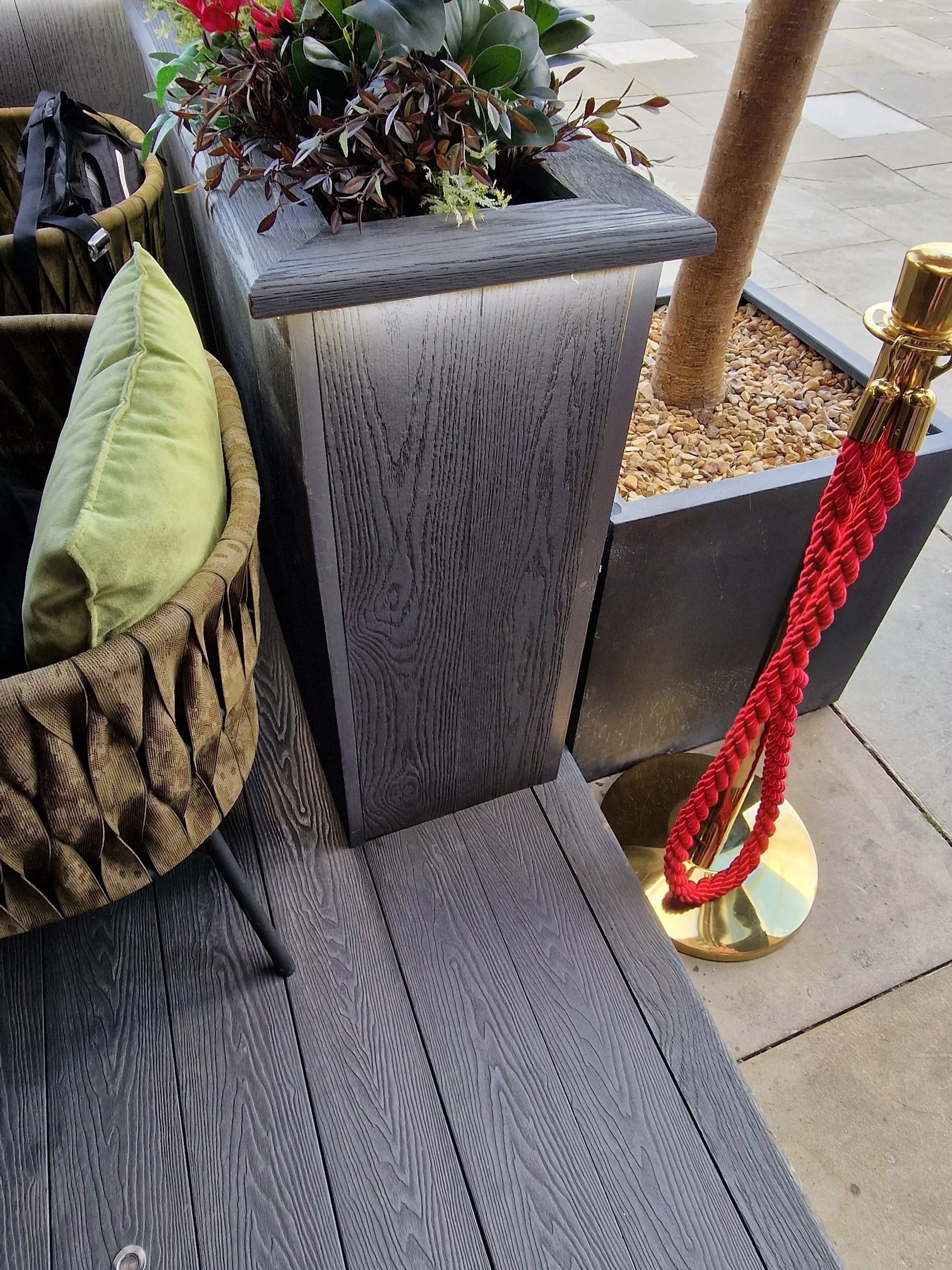 A wooden bench with black finish next to a large planter box with green and red plants, a yellow pillow on a woven chair, and a red rope barrier with gold posts outside on a tiled sidewalk.