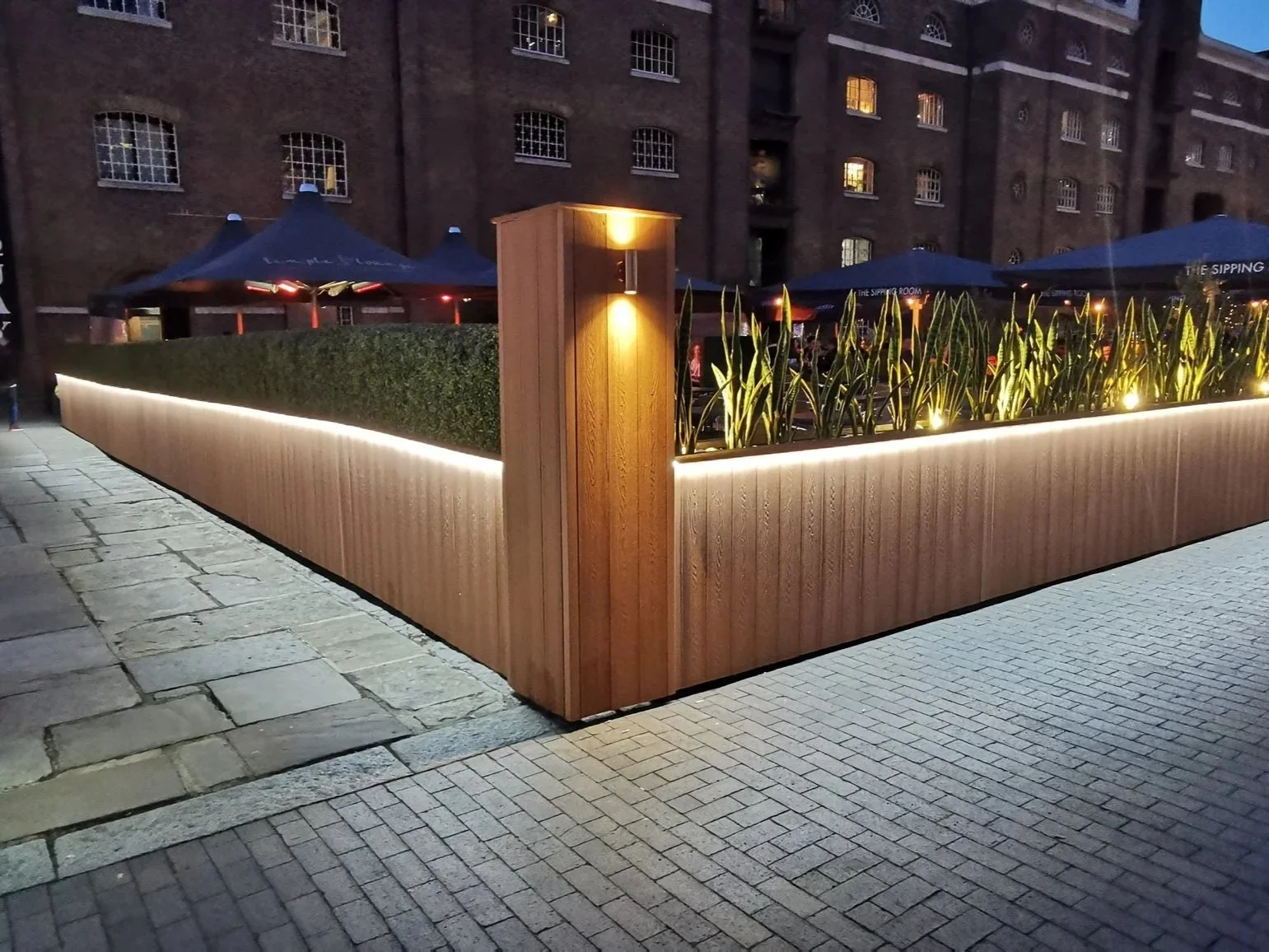 An outdoor elevated planter with green plants, illuminated by lighting along its base, situated on a brick sidewalk in front of a brick building with windows and outdoor seating with umbrellas at dusk.
