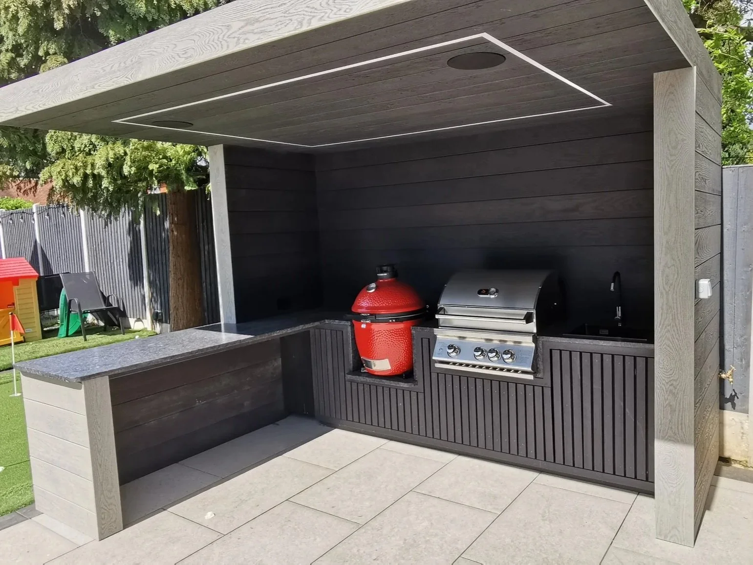 An outdoor backyard barbecue area with a built-in grill, red smoker, and countertop, surrounded by a children's play area with a small yellow playhouse and toys, situated on a lawn with trees and a fence in the background.