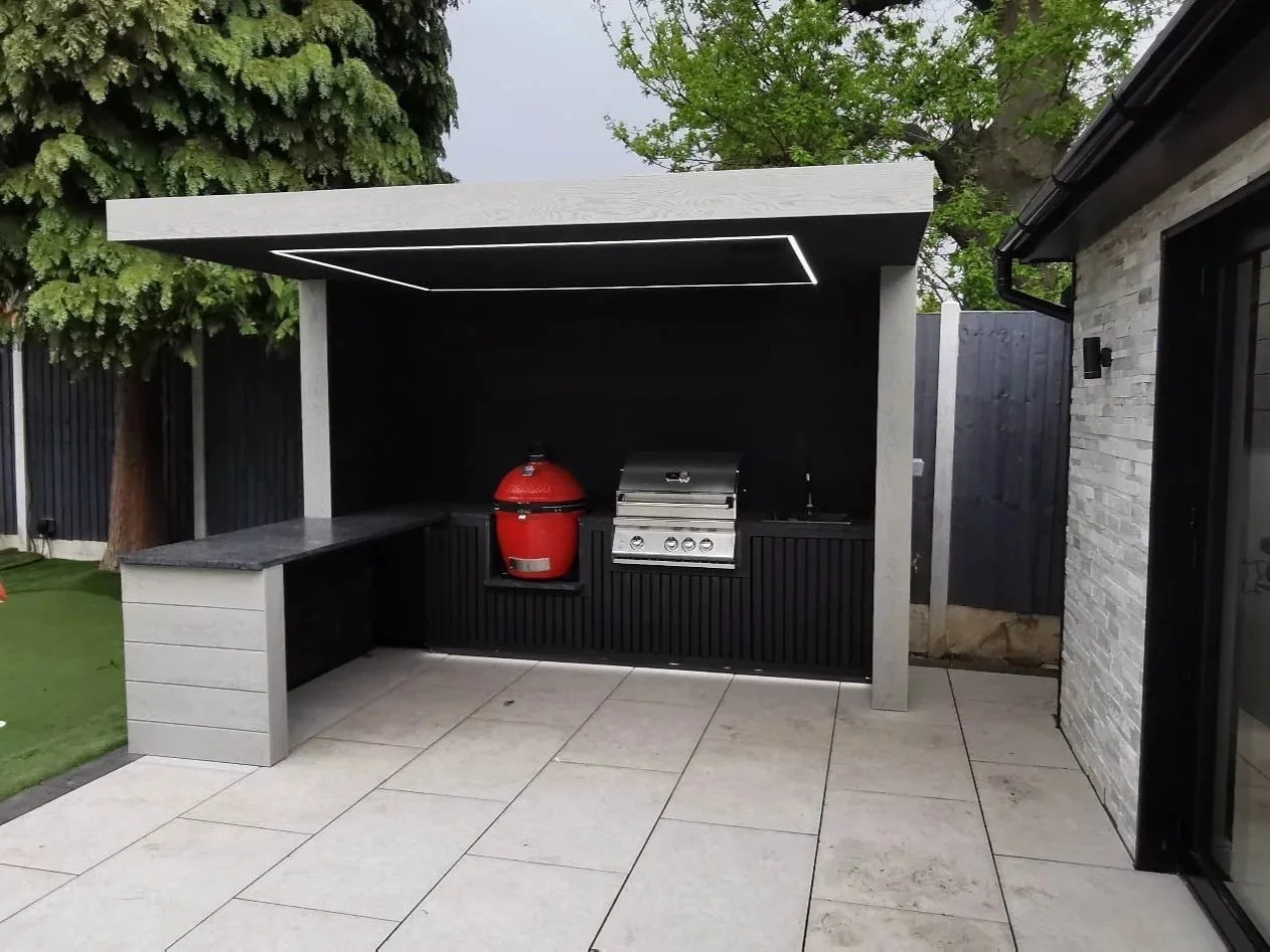Outdoor kitchen area with a built-in grill, red smoker, and countertop, surrounded by a black wall and modern design elements. Outdoor kitchen