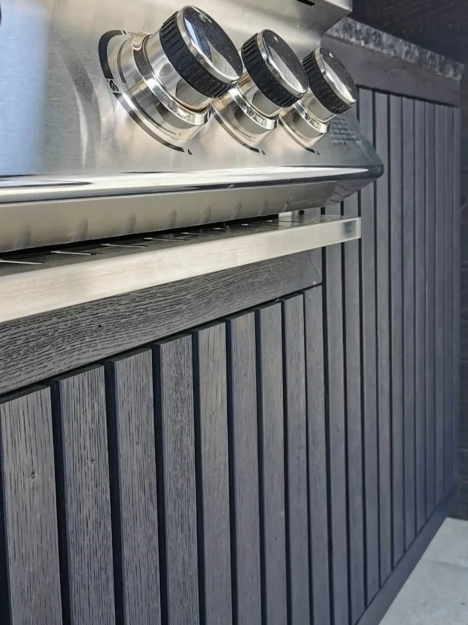 Close-up of stainless steel grill with three black control knobs mounted on a dark wood-paneled exterior wall.
