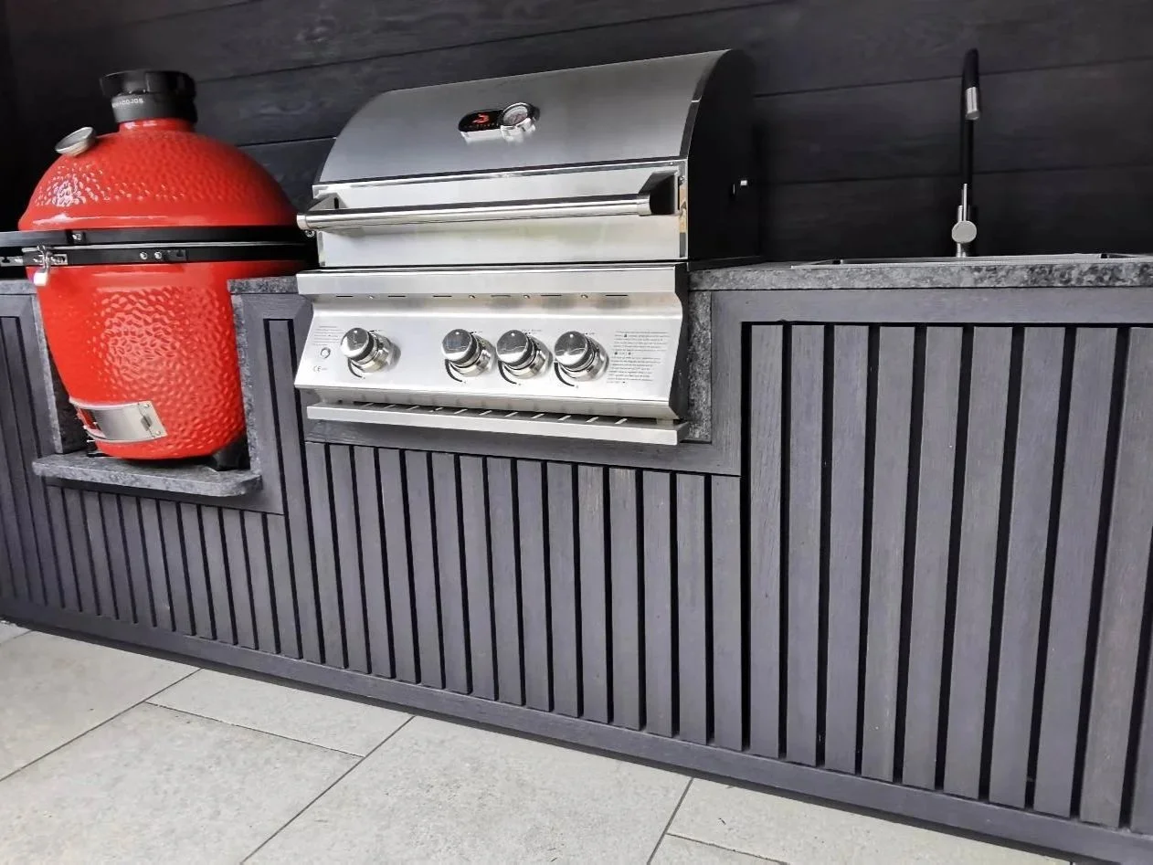A red smoker grill and a stainless steel gas grill on a black wooden outdoor kitchen counter with a sink.