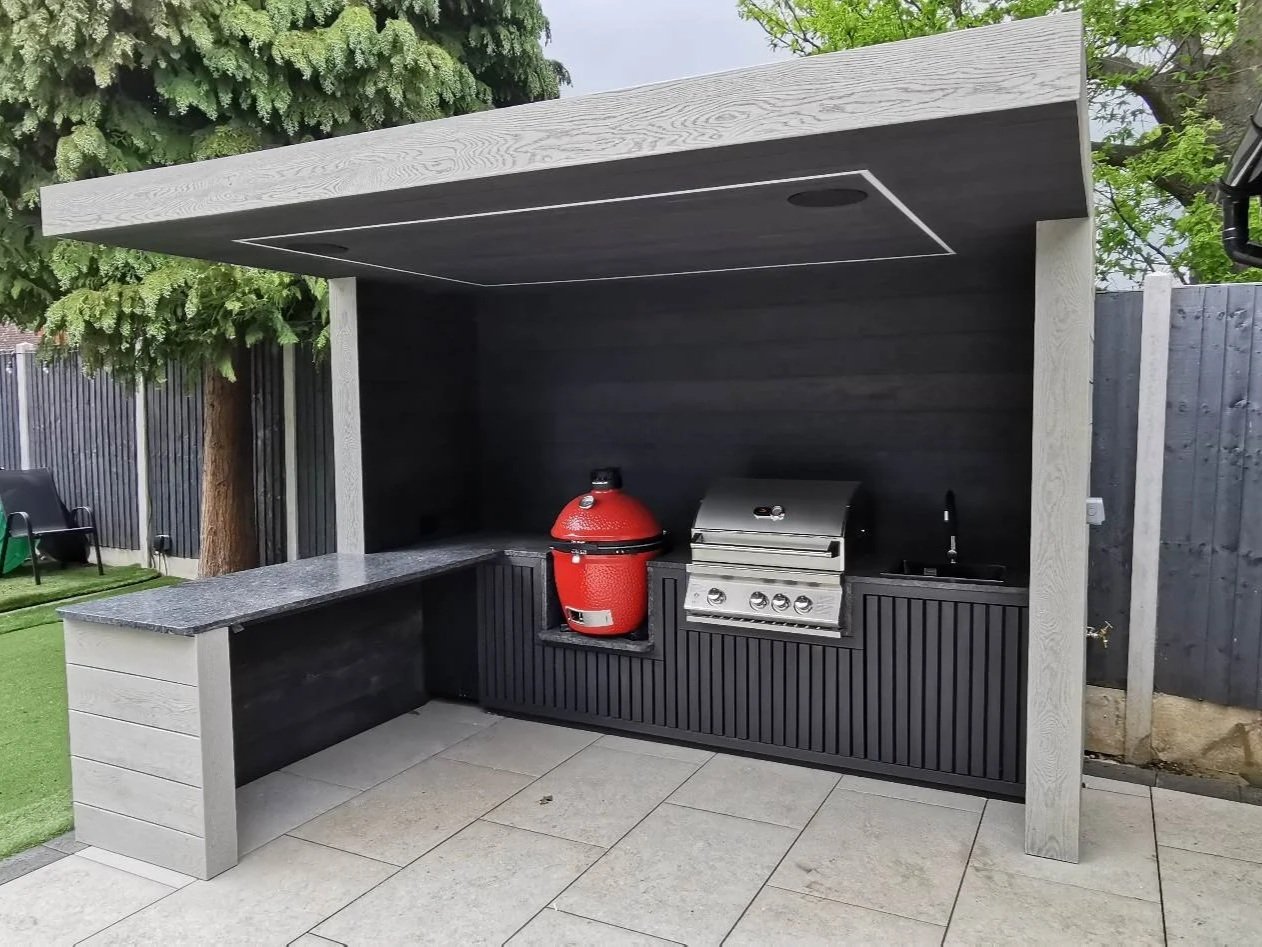 Outdoor kitchen with a built-in grill, red smoker, and sink, under a modern black and gray canopy with a tiled countertop.