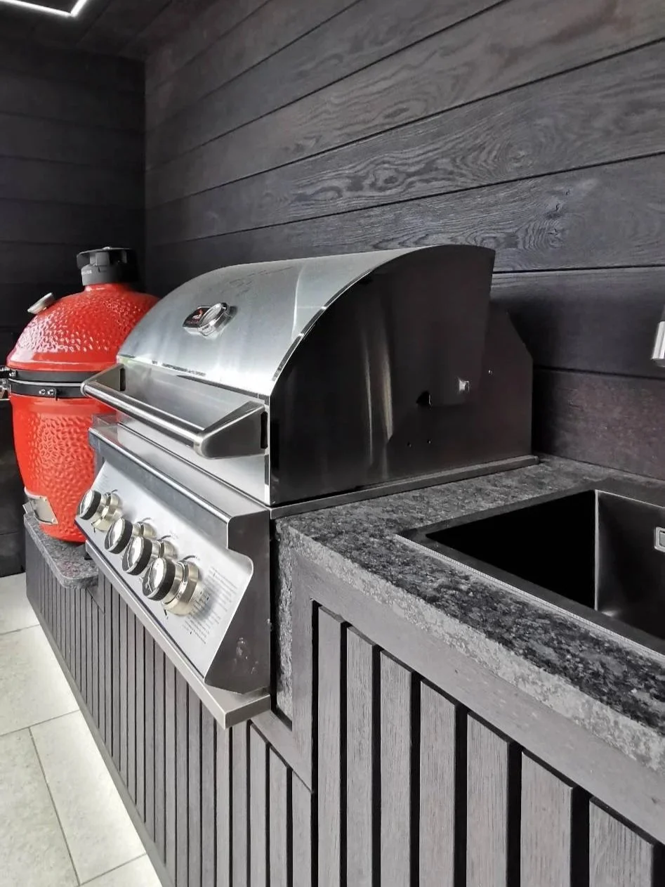 A stainless steel outdoor grill on a dark stone countertop next to a red ceramic grill and dark wood paneling.