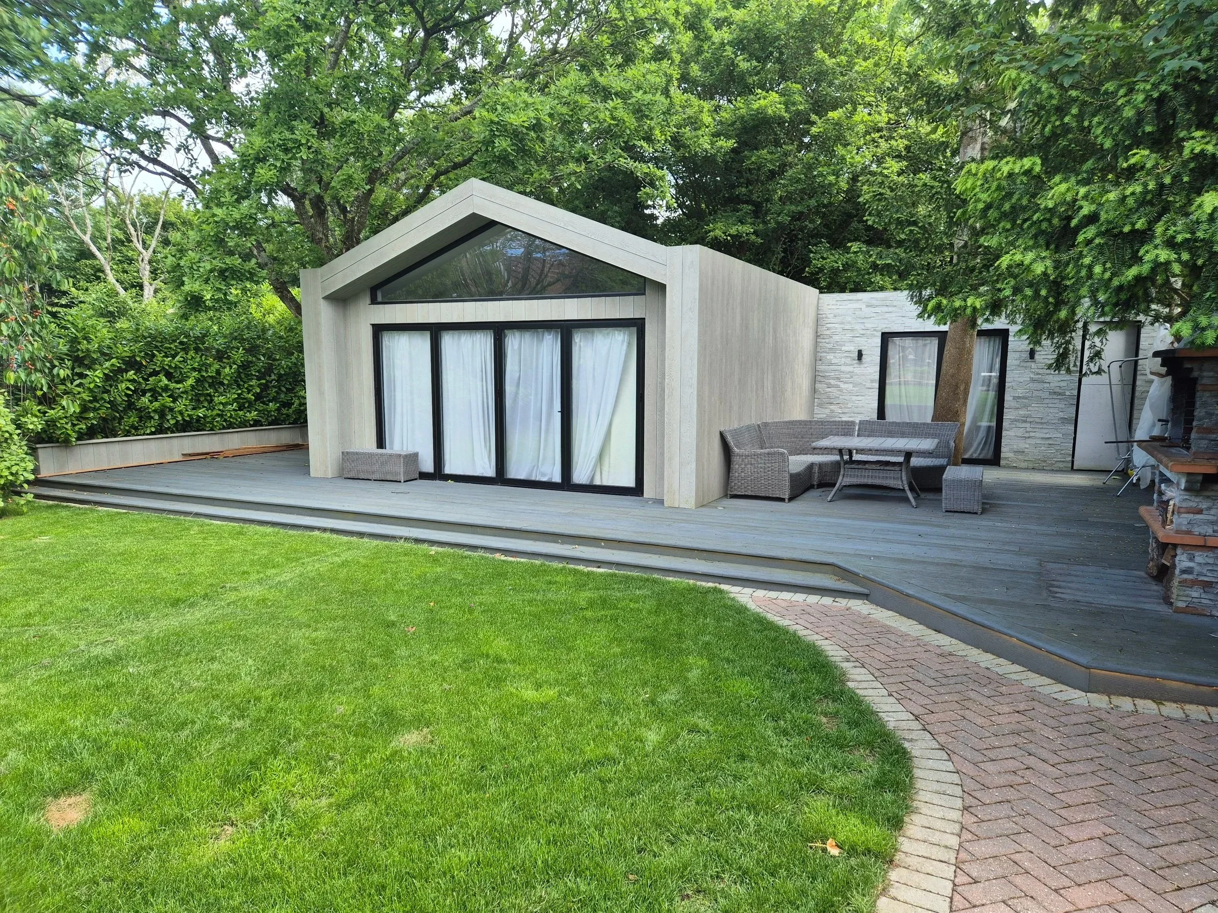 A modern house with large glass doors and windows, set in a lush garden with mature trees, a wooden deck, and outdoor seating.