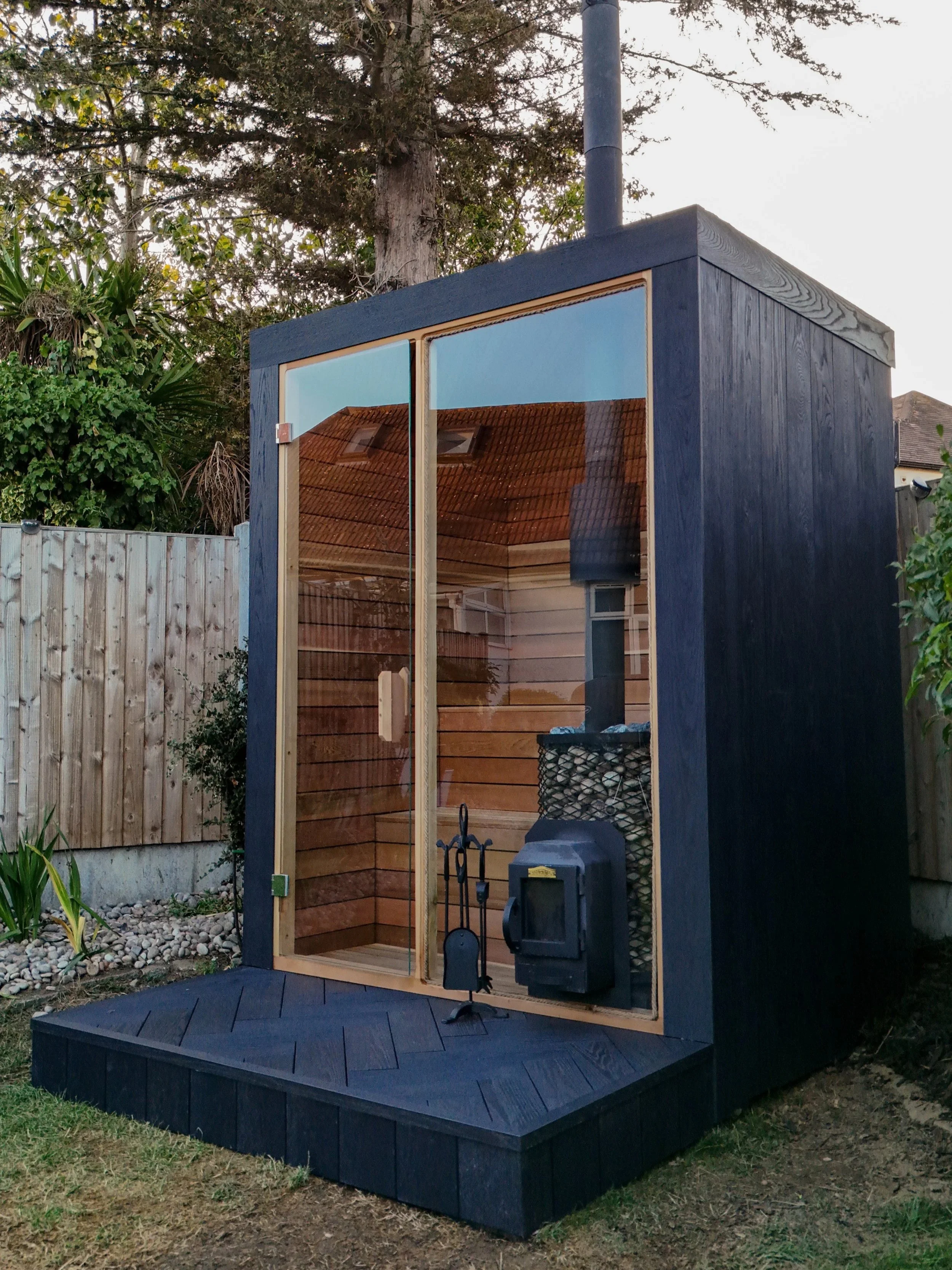 A small, modern outdoor wooden and glass structure with a black exterior and large glass door, set in a backyard with trees, a wooden fence, and some garden plants.