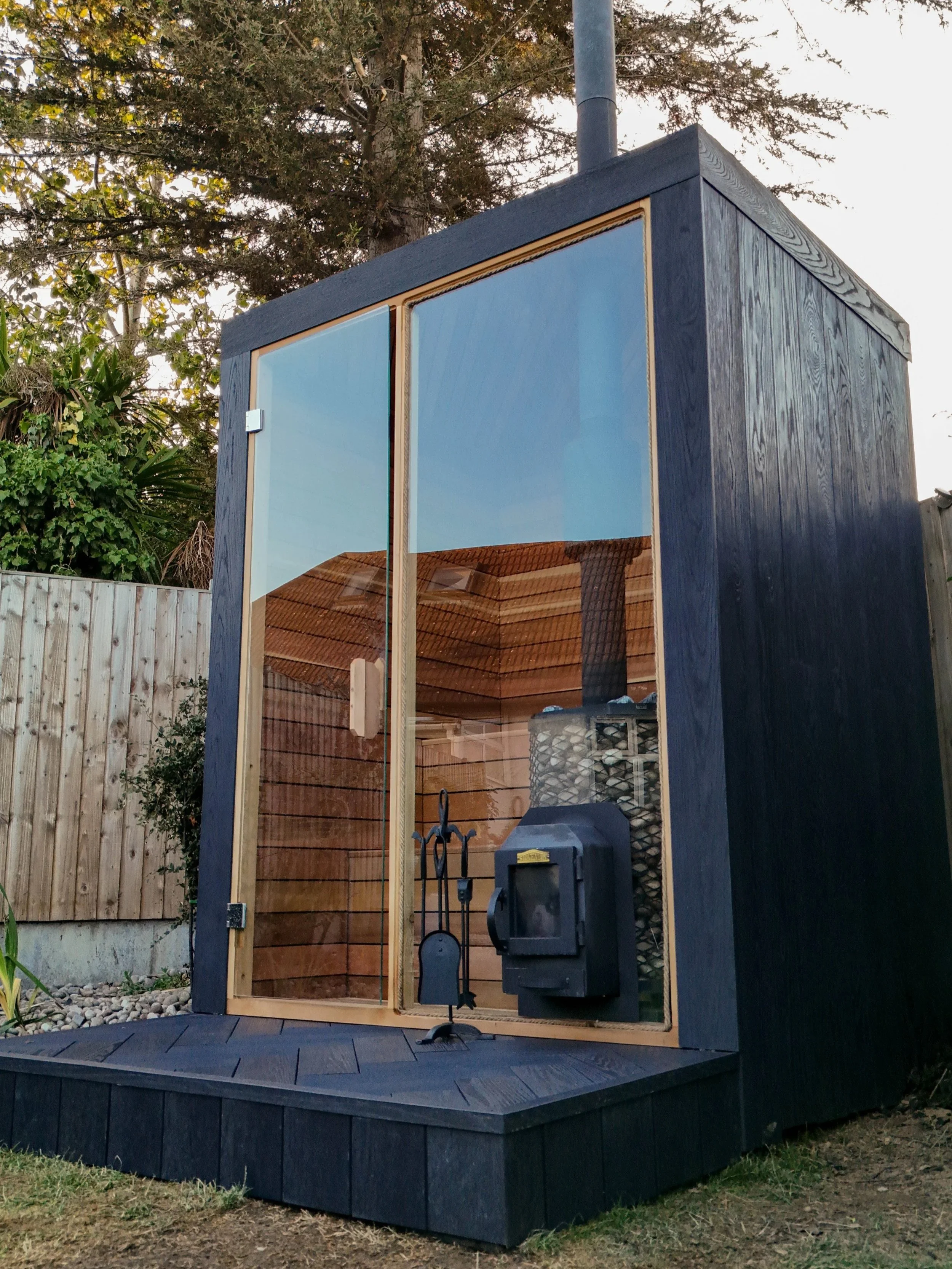 A modern outdoor sauna made of dark wood with large glass windows, situated in a backyard with a wooden fence and trees in the background.