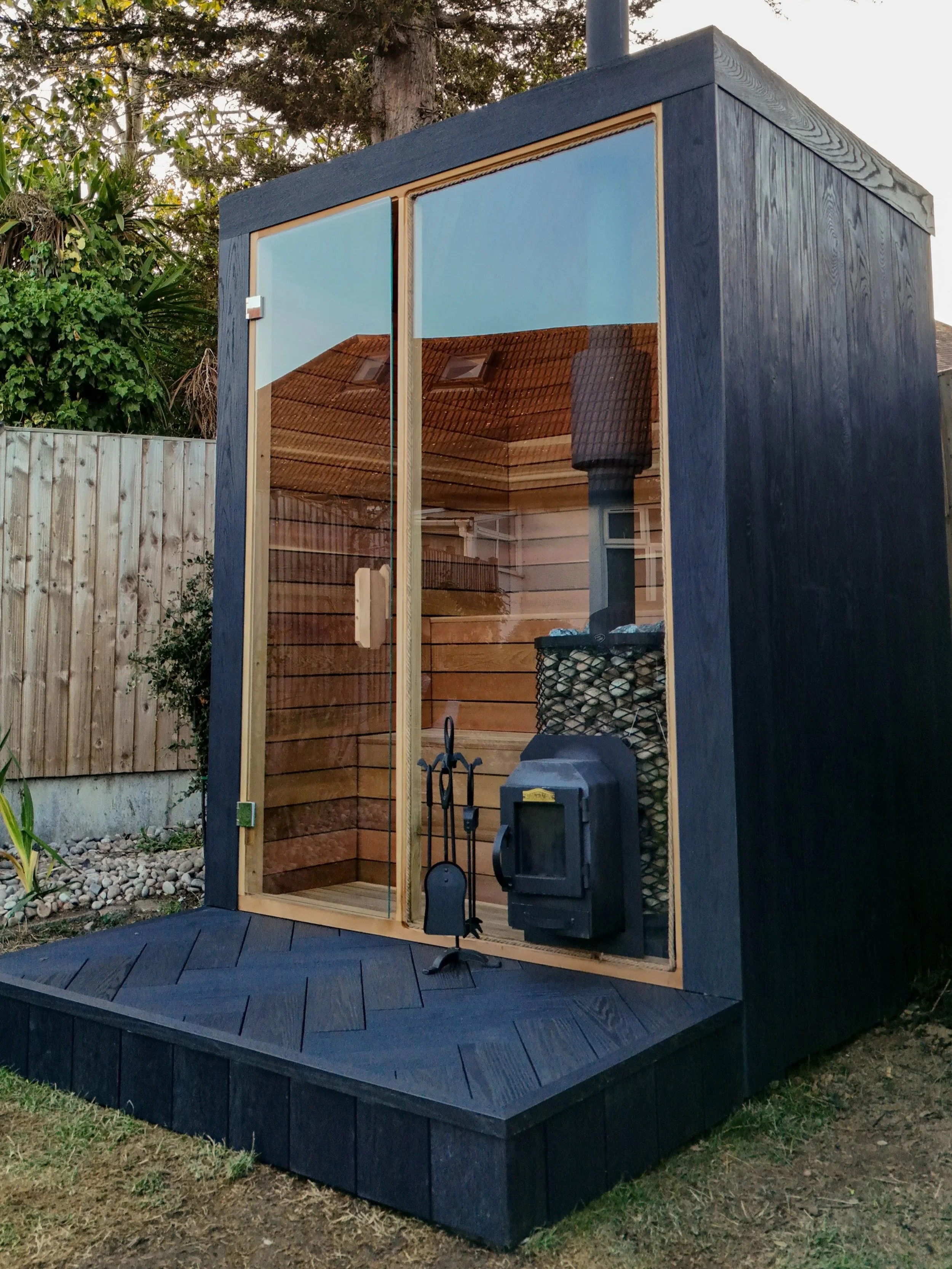 A modern outdoor sauna with a black exterior and large glass door, situated on a wooden deck in a backyard with trees and a wooden fence.