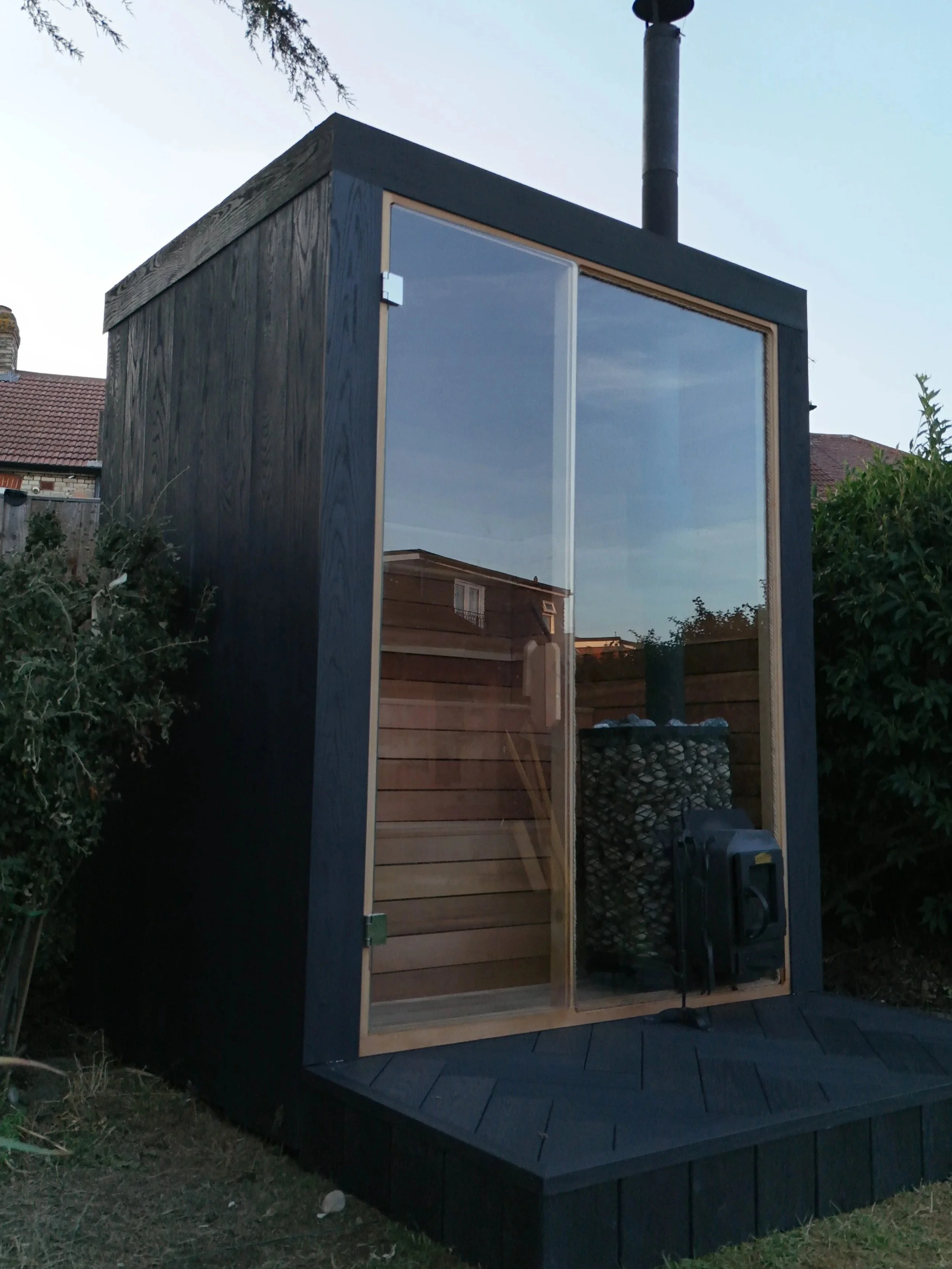 A modern outdoor sauna with a black exterior, large glass front door, and a wood interior, situated in a garden area with plants and neighboring houses visible.