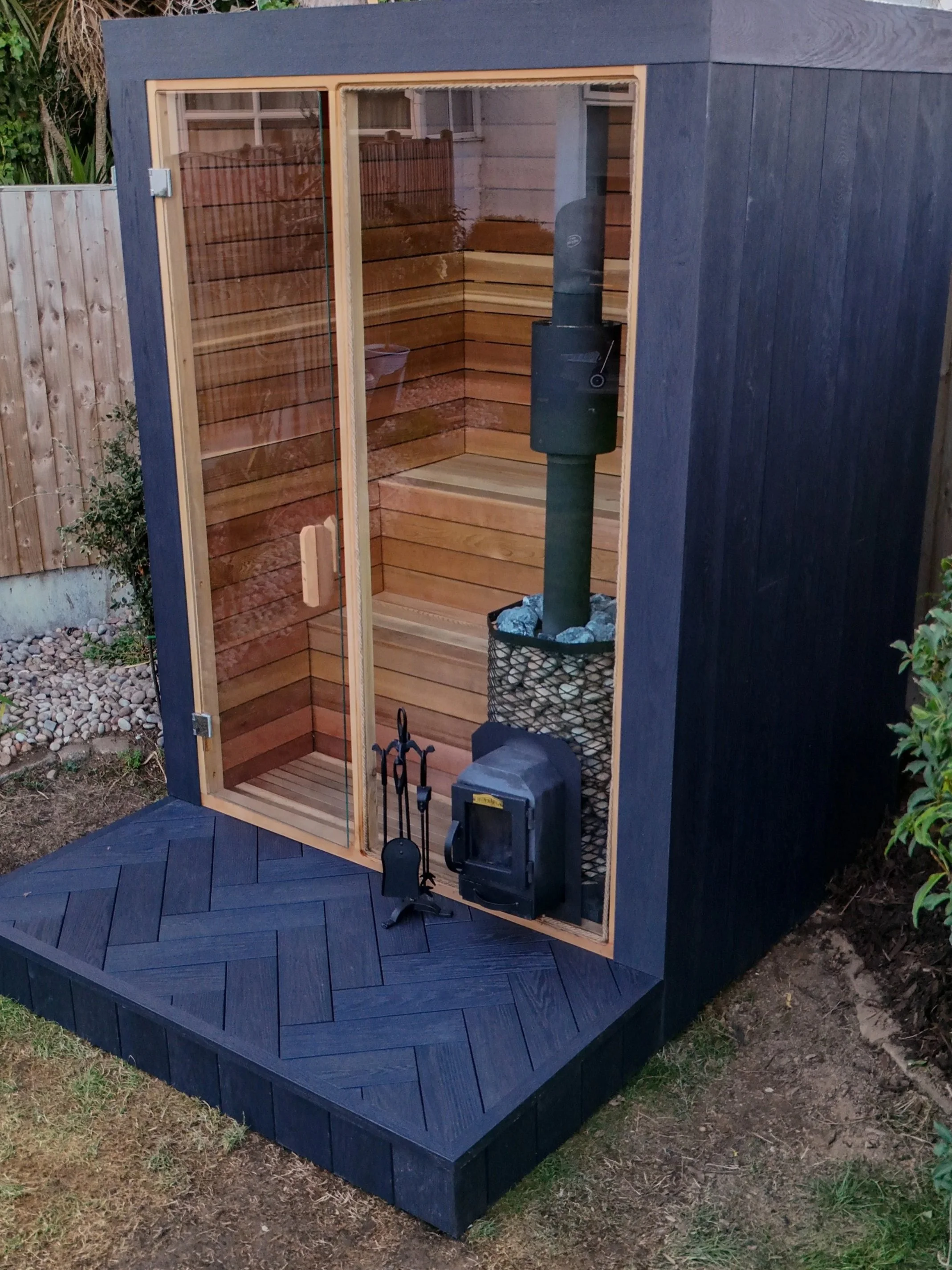 A small outdoor sauna shed with a glass door, wooden interior, and a stove with rocks inside for heating, situated on a deck with a black painted exterior.
