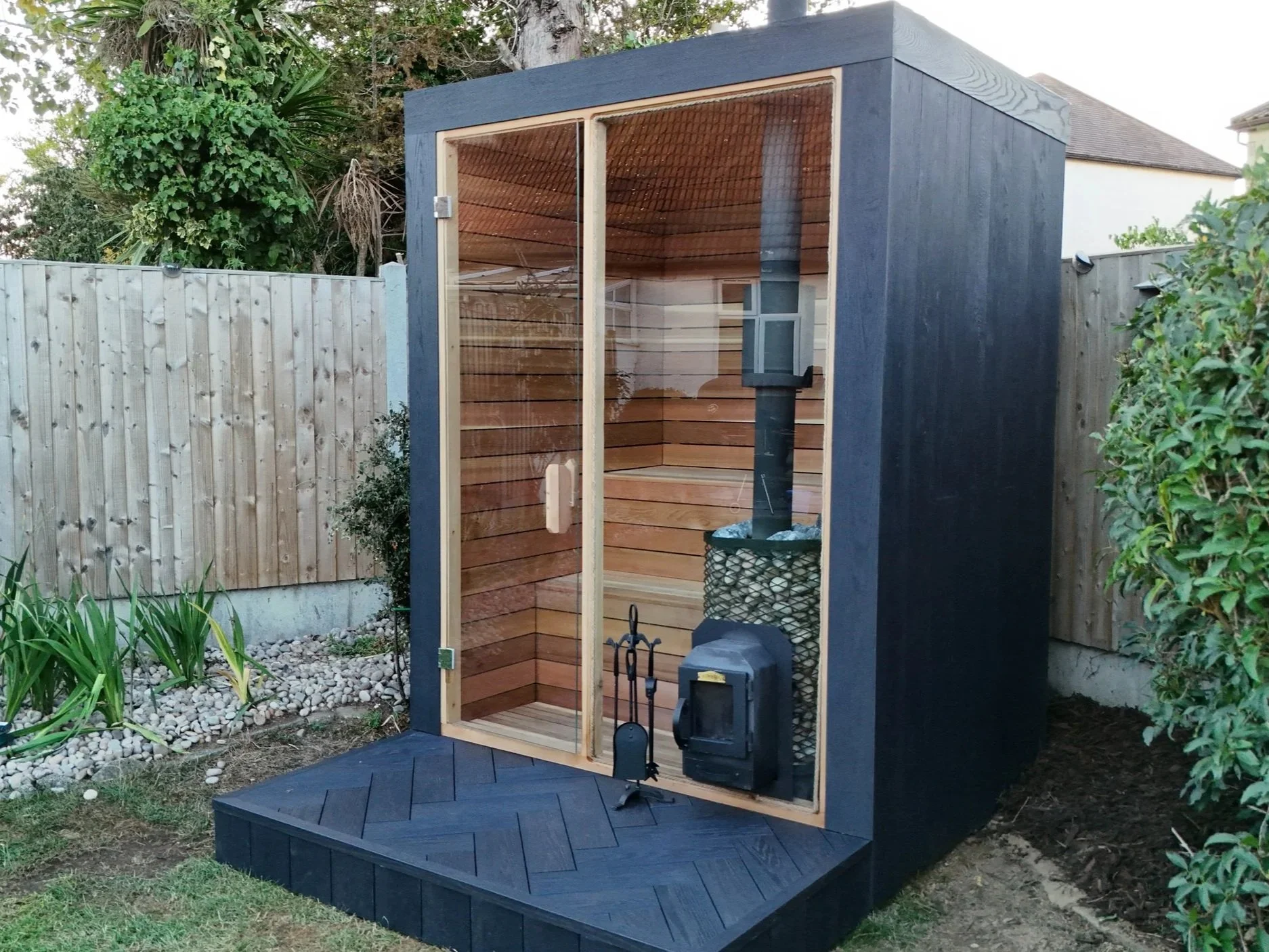 Small black outdoor sauna with large glass door, interior shows wood paneling and sauna stove, situated in a garden with plants, trees, and a wooden fence.