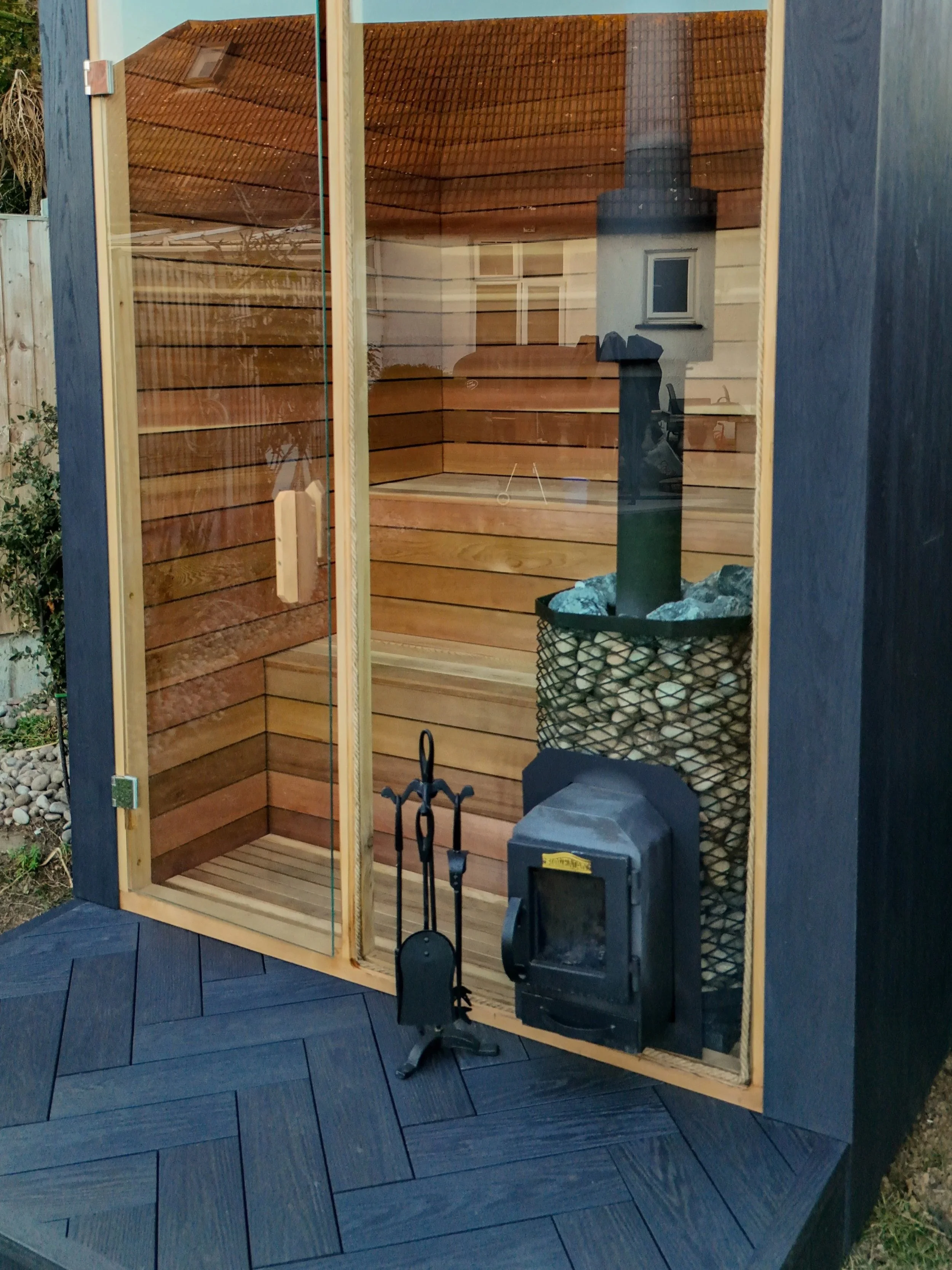 Outdoor sauna with glass door, wooden interior with benches, heater with stones, and black tools hanging outside on a dark wood deck.