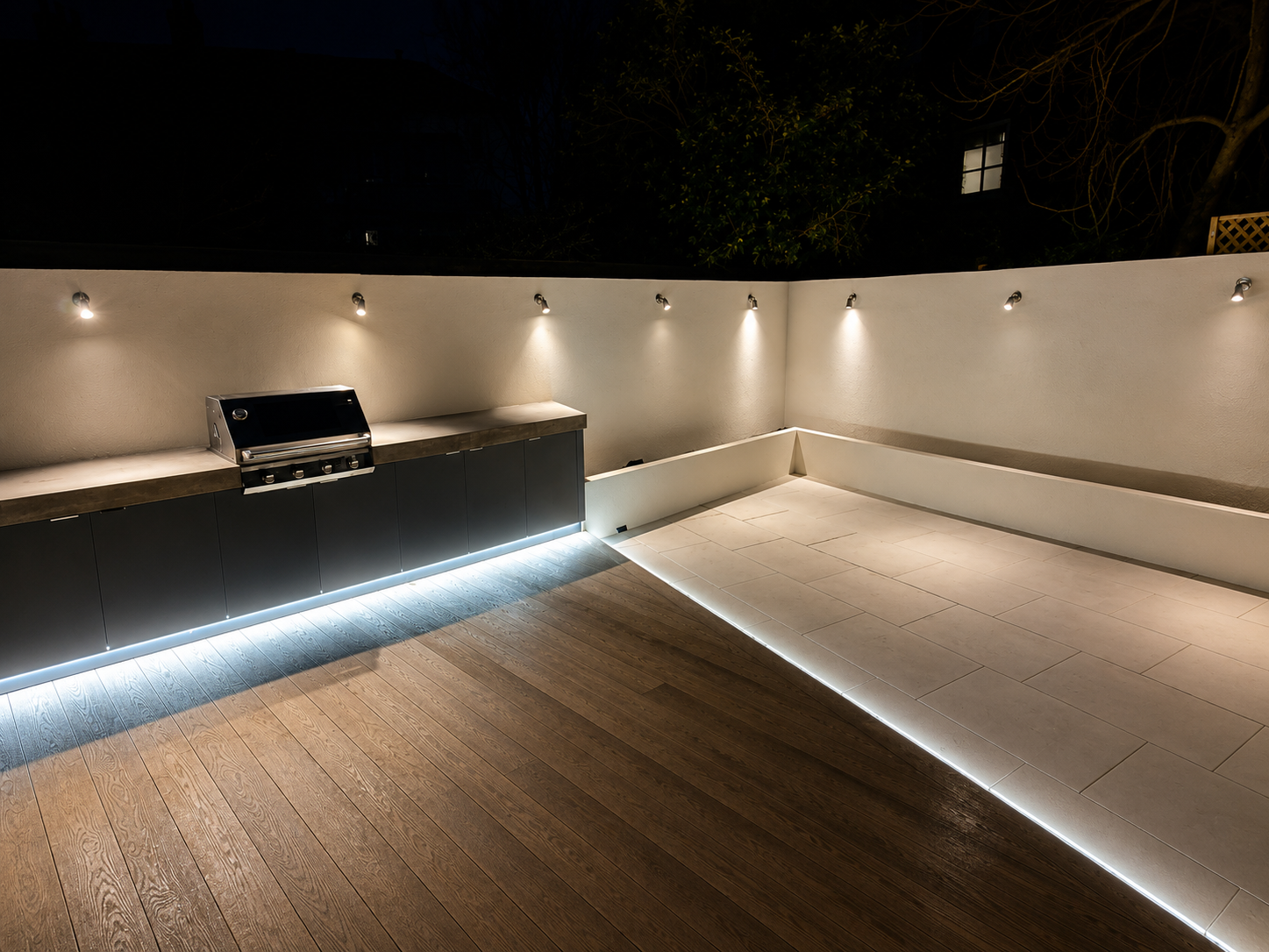 Outdoor kitchen area with built-in grill, modern lighting, and wooden and tiled flooring at night.