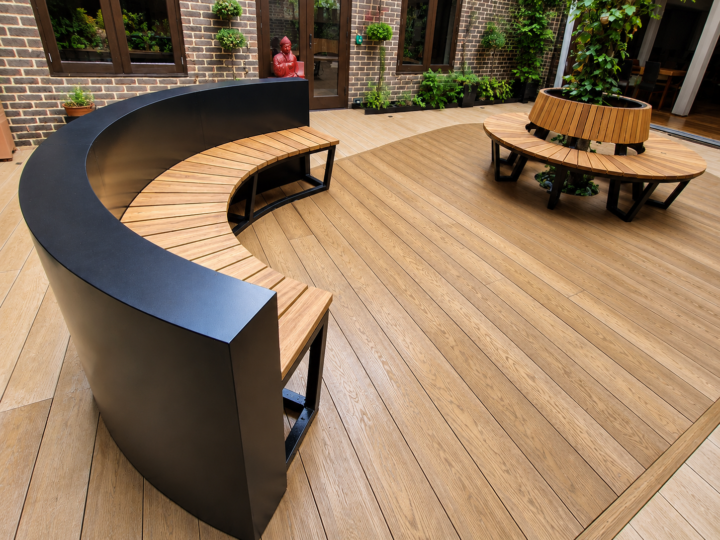 Outdoor patio with a curved wooden bench with black backing and a separate circular wooden plant bed, surrounded by potted plants and brick walls.