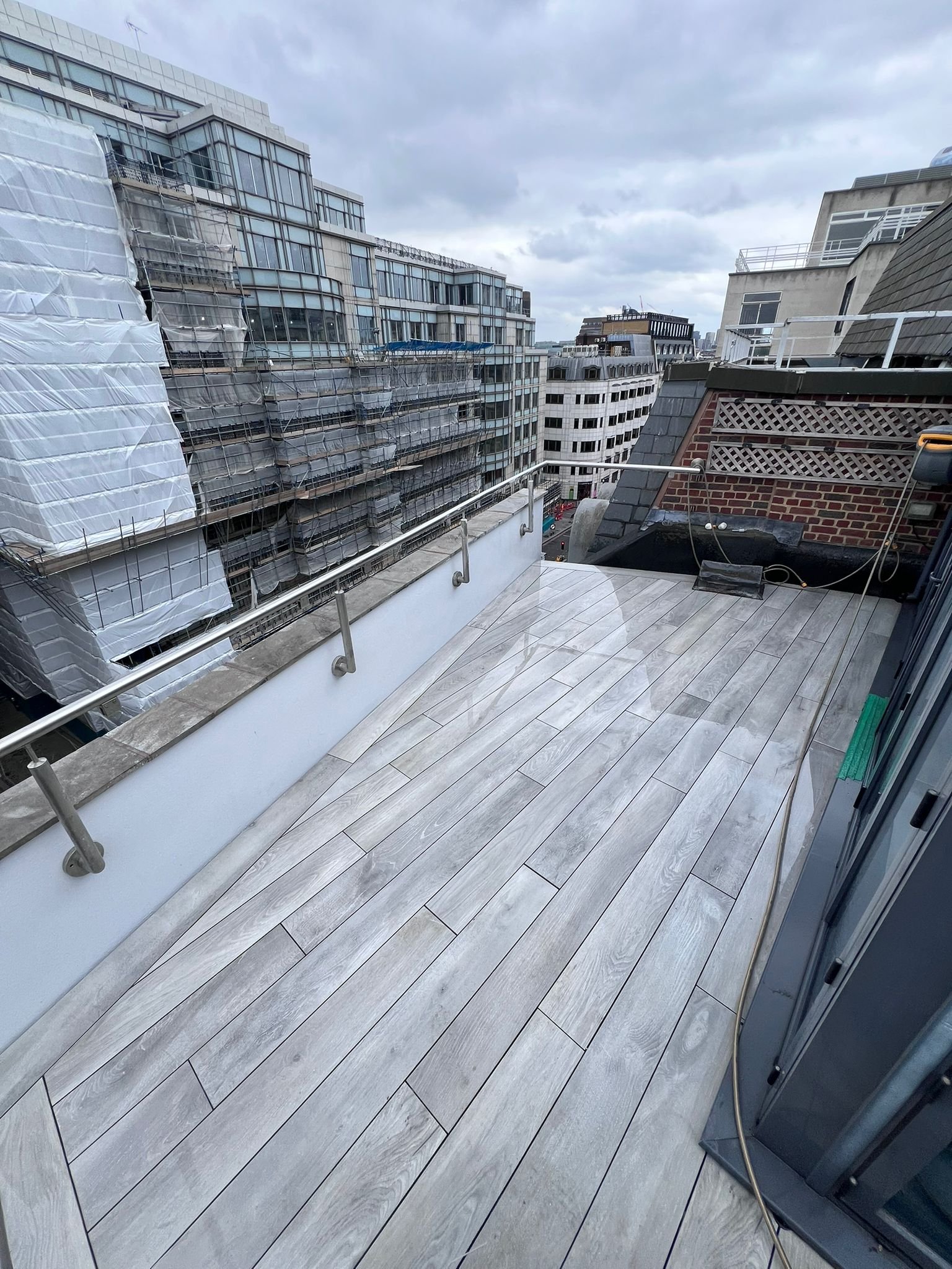 A rooftop terrace area with light-colored wood plank flooring, a metal railing, and neighboring high-rise buildings with glass windows and modern architecture under a cloudy sky.
