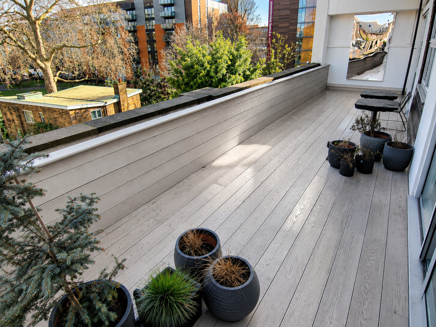 Empty balcony with potted plants, black chairs, wooden flooring, and city buildings in the background.