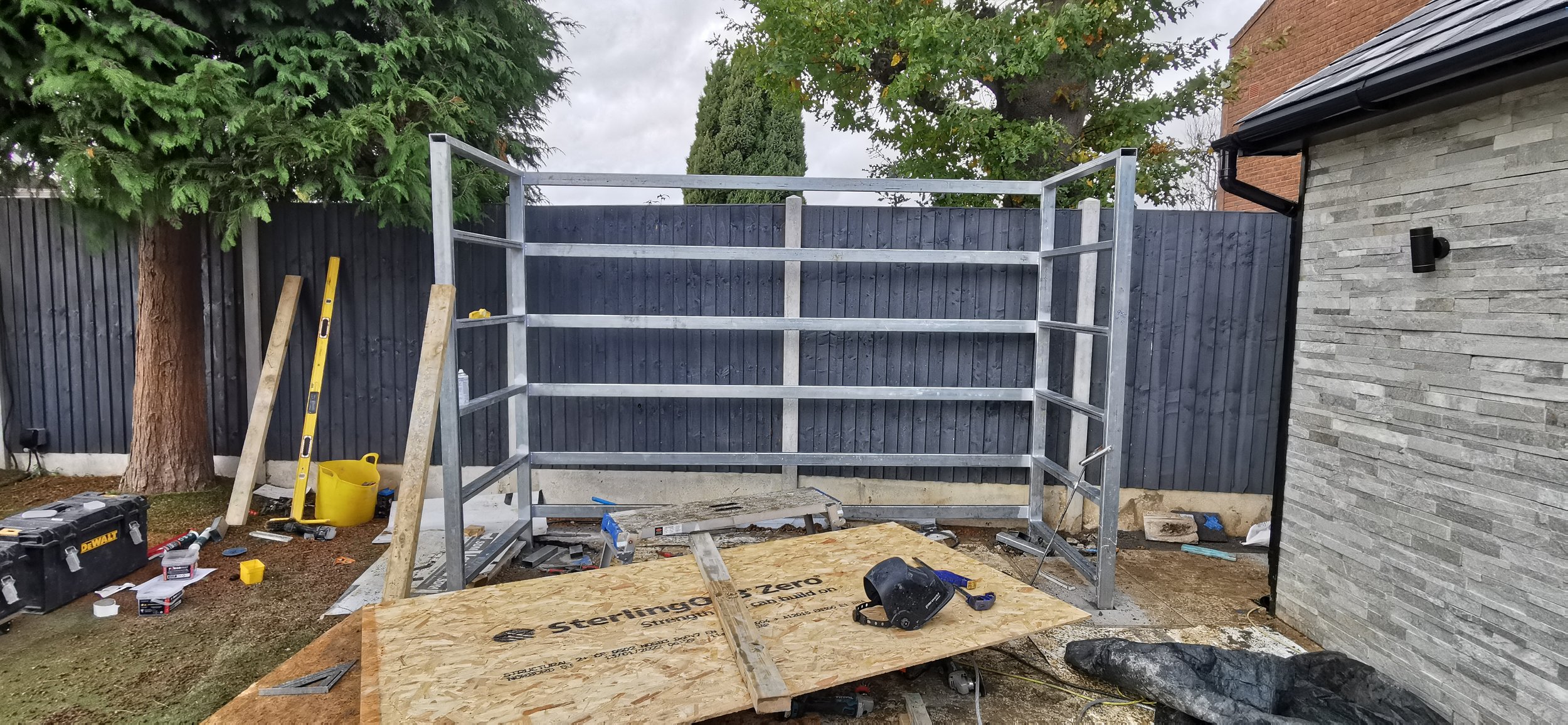 A partially assembled metal outdoor shelf unit in a backyard with construction tools and materials scattered around, next to a tree, a wooden fence, and a house wall.