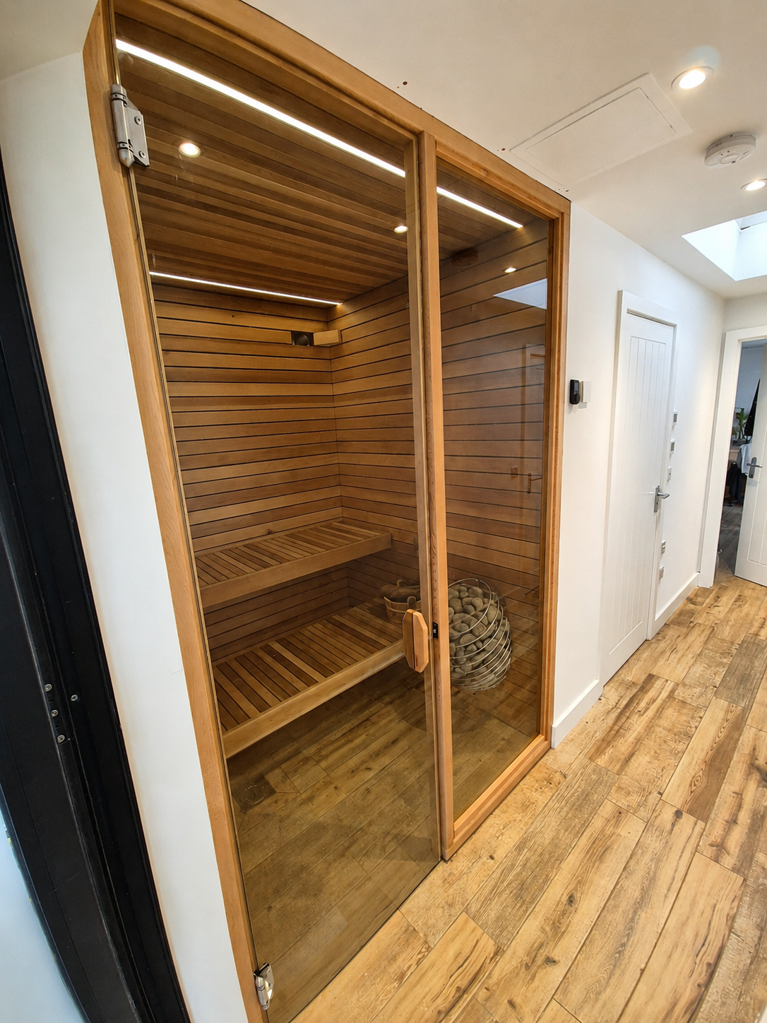 Indoor wooden sauna with glass door, wooden benches inside, and sauna stones in a metal heater.