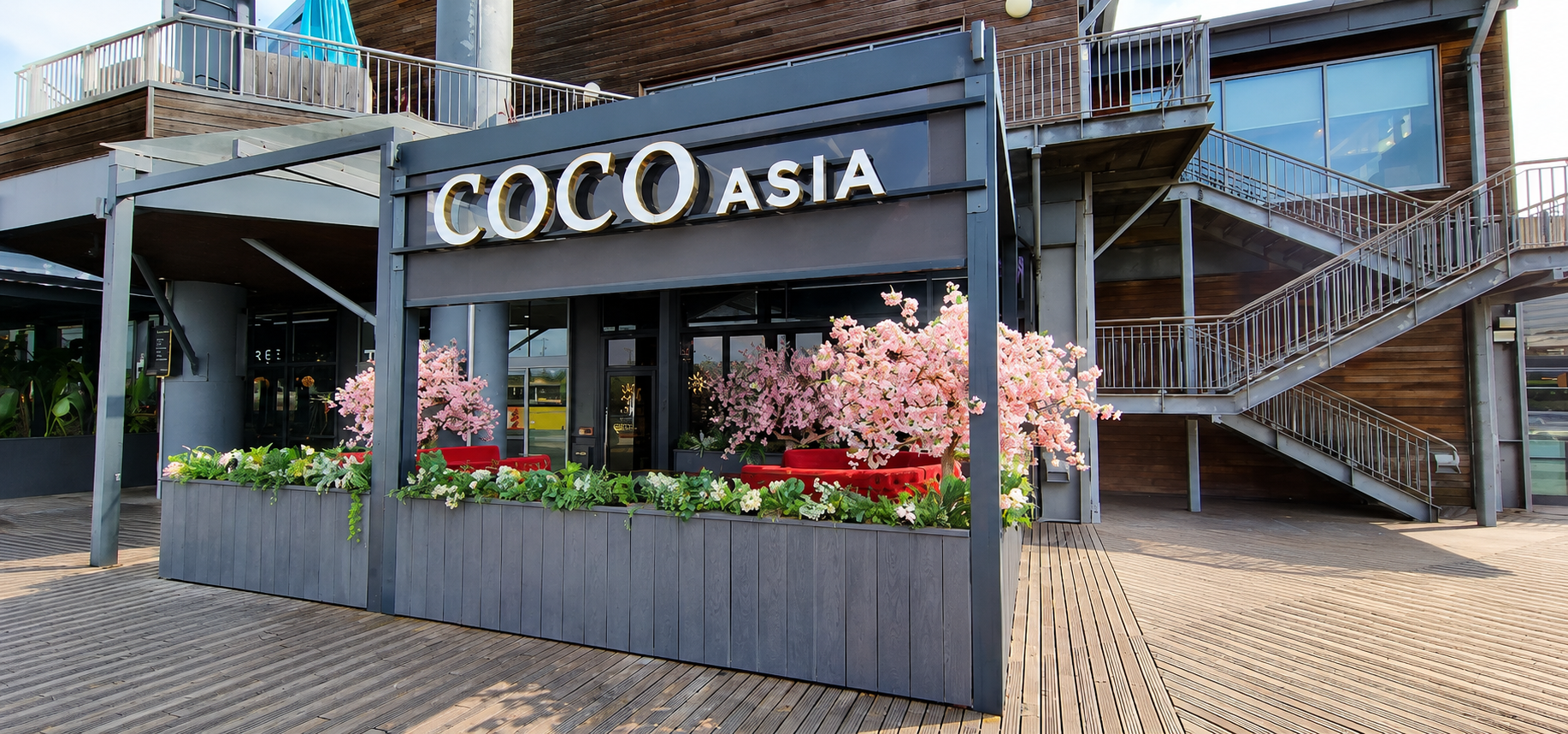 Exterior of a modern building with a sign that says 'COCO ASIA', pink flowering trees, and outdoor seating with red cushions on a wooden deck.
