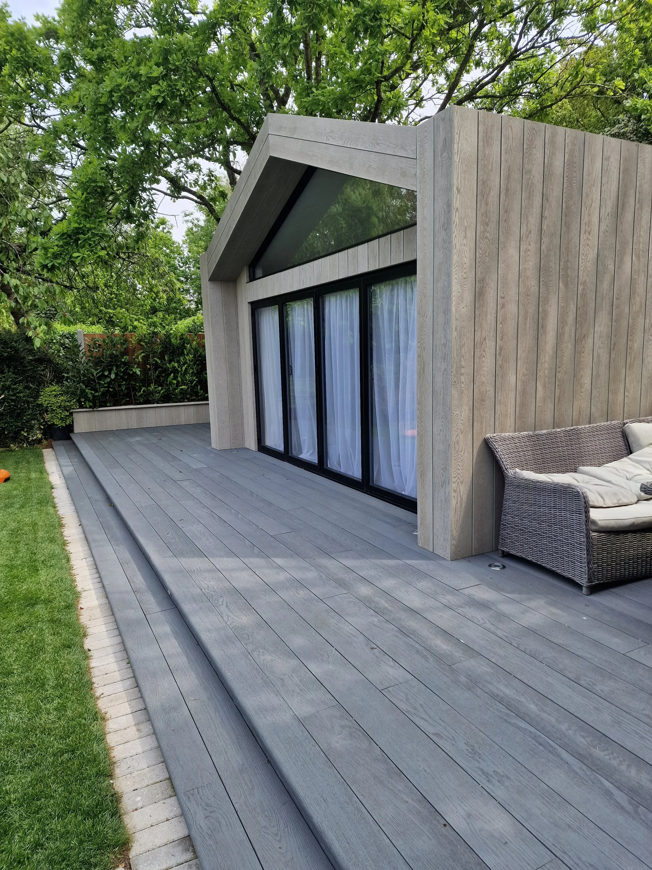 Modern house with large glass doors and wooden exterior, located in a lush green backyard with trees and a patio with outdoor furniture.