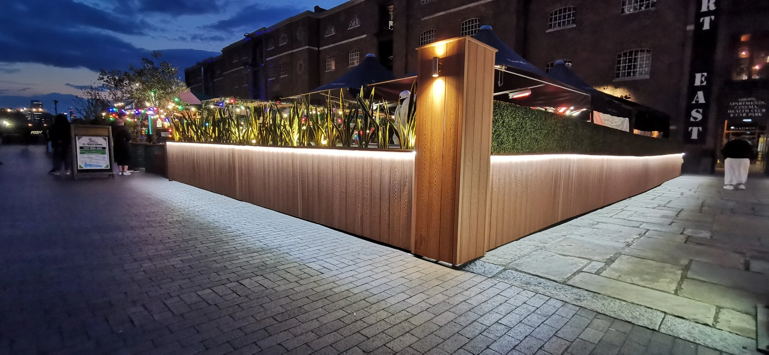 Night view of an outdoor dining area with a wooden fence, plants, and warm lighting, with a brick building and city lights in the background.
