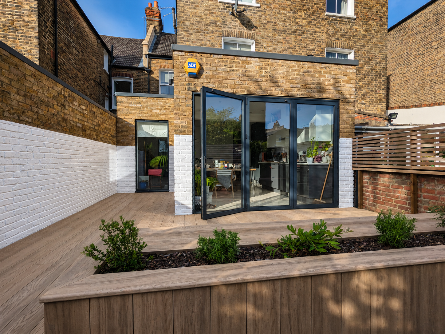Backyard patio with wooden deck, white-painted brick wall, planters with green plants, and a glass sliding door leading into a house with brick exterior.
