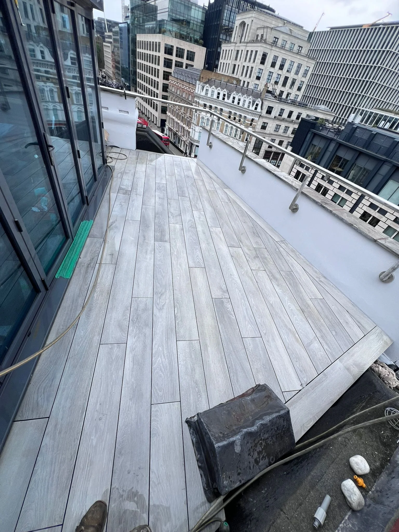 Newly installed light-colored wooden flooring on a city balcony with metal railing, surrounded by tall office buildings. Construction tools and materials are visible at the bottom of the balcony.