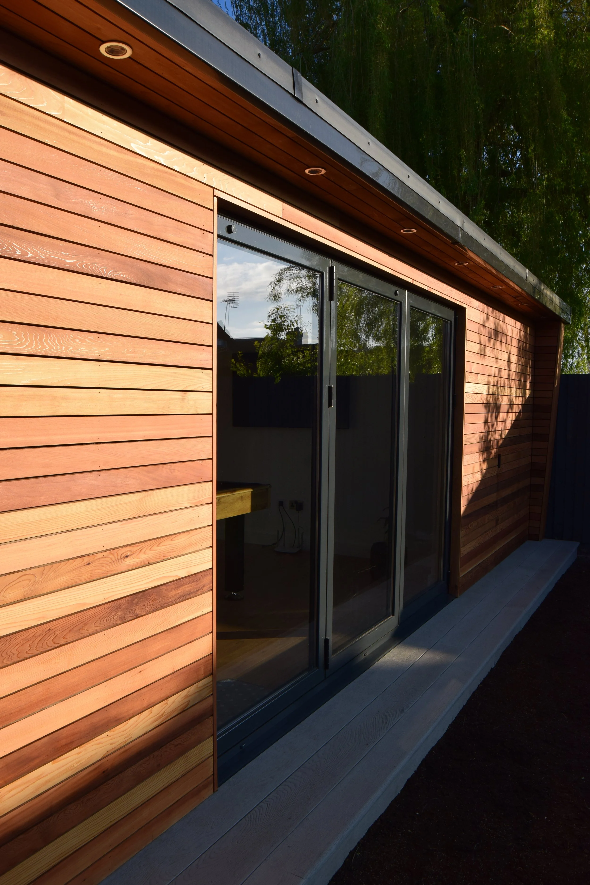 Close-up view of a modern house exterior with reddish-brown wooden siding, dark metal trim, and large sliding glass doors reflecting trees and sky. The house has a deck with gray wooden planks.