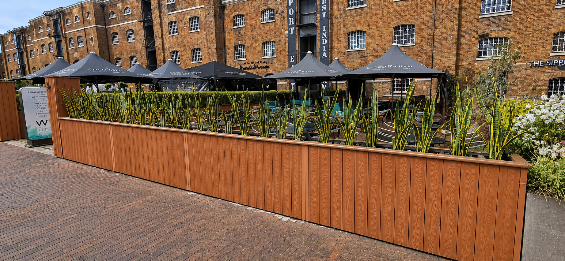 Outdoor patio with tables, chairs, and black umbrellas, enclosed by a large wooden planter filled with tall green plants, in front of a brick building with several windows.