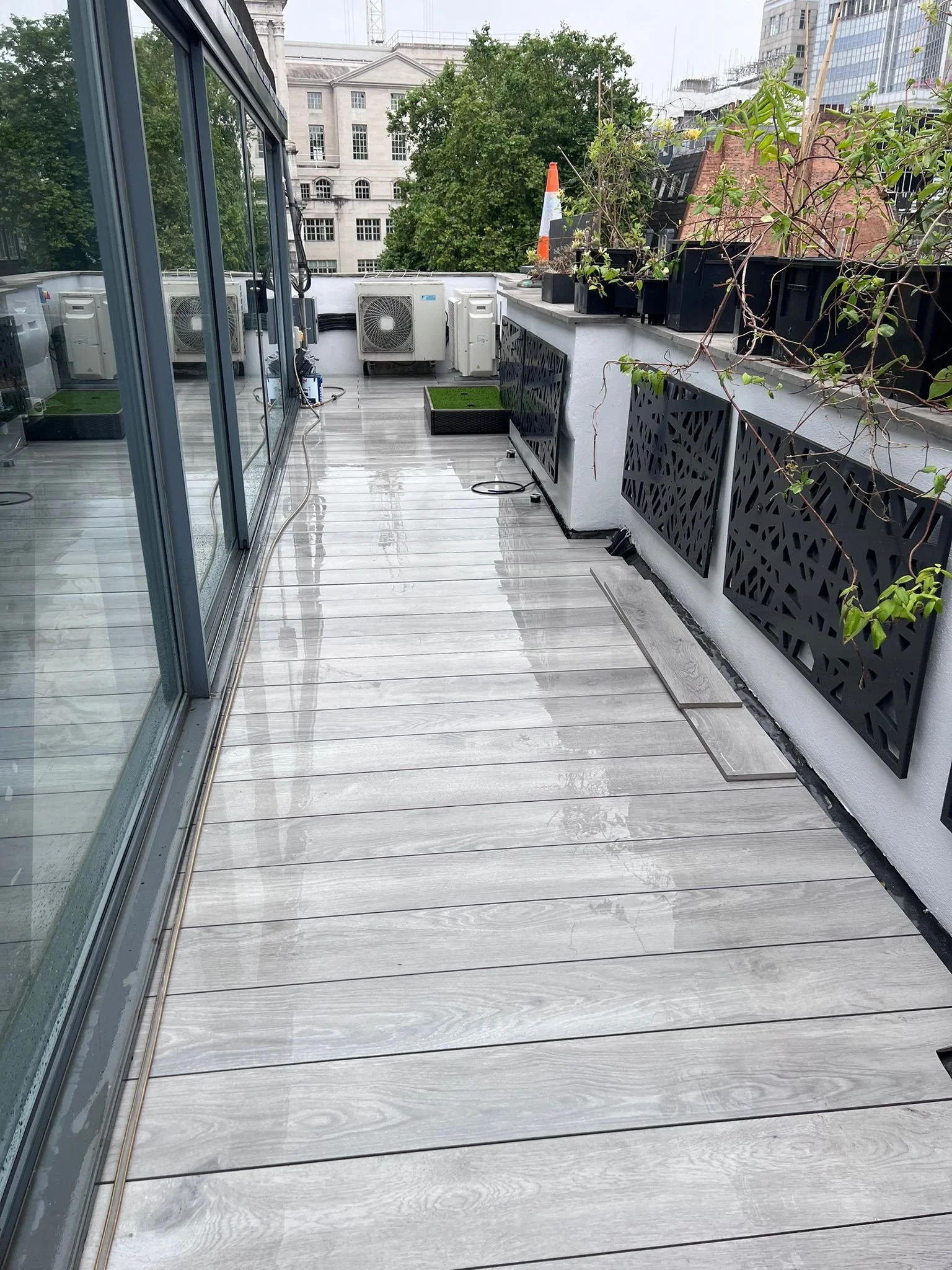 View of a modern balcony with light-colored wooden flooring, black decorative panels on the wall, potted plants, and city buildings in the background.