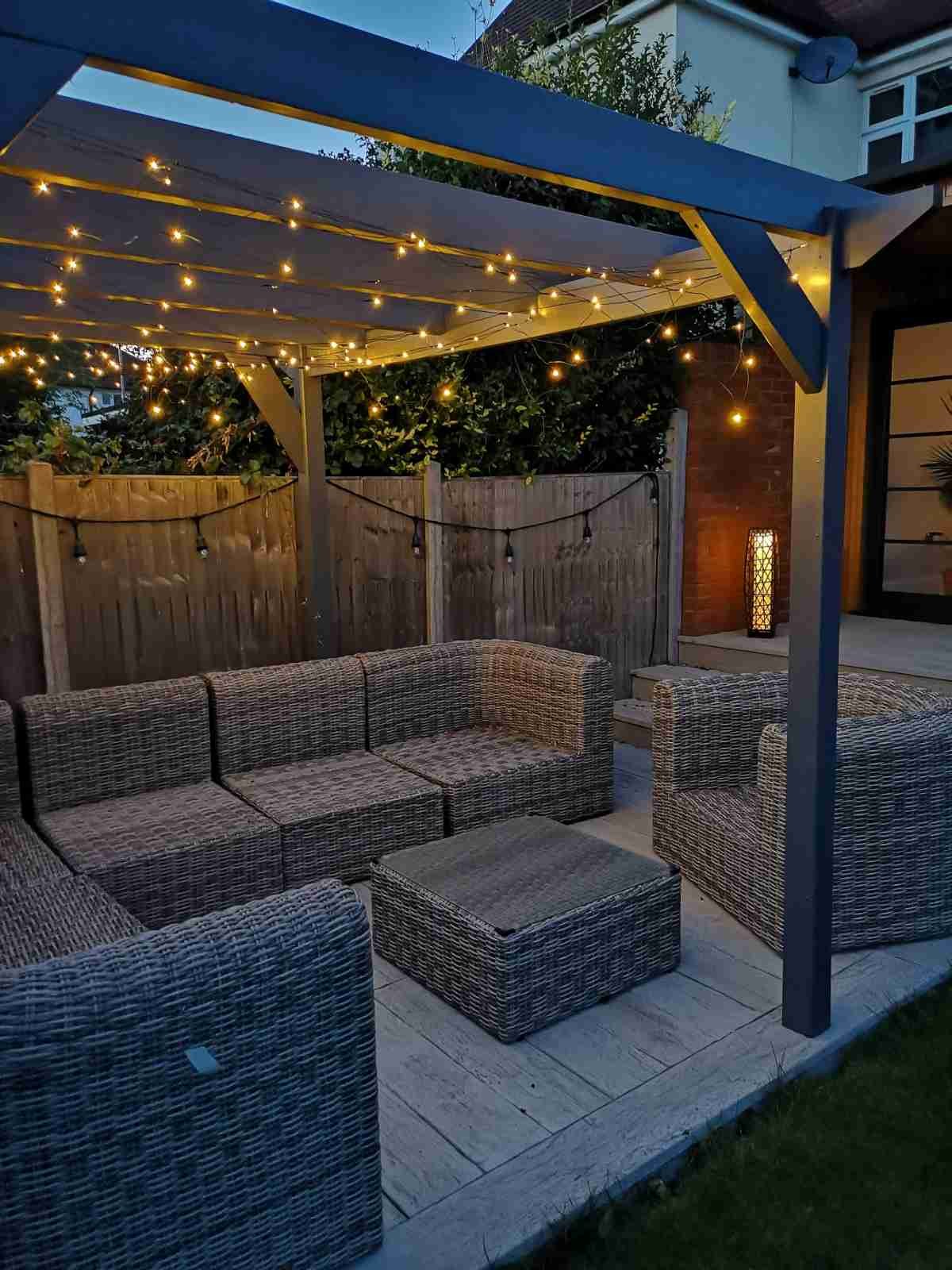 Outdoor patio with wicker furniture, string lights hanging under a wooden pergola, a wooden fence, and a brick wall with a decorative lamp.
