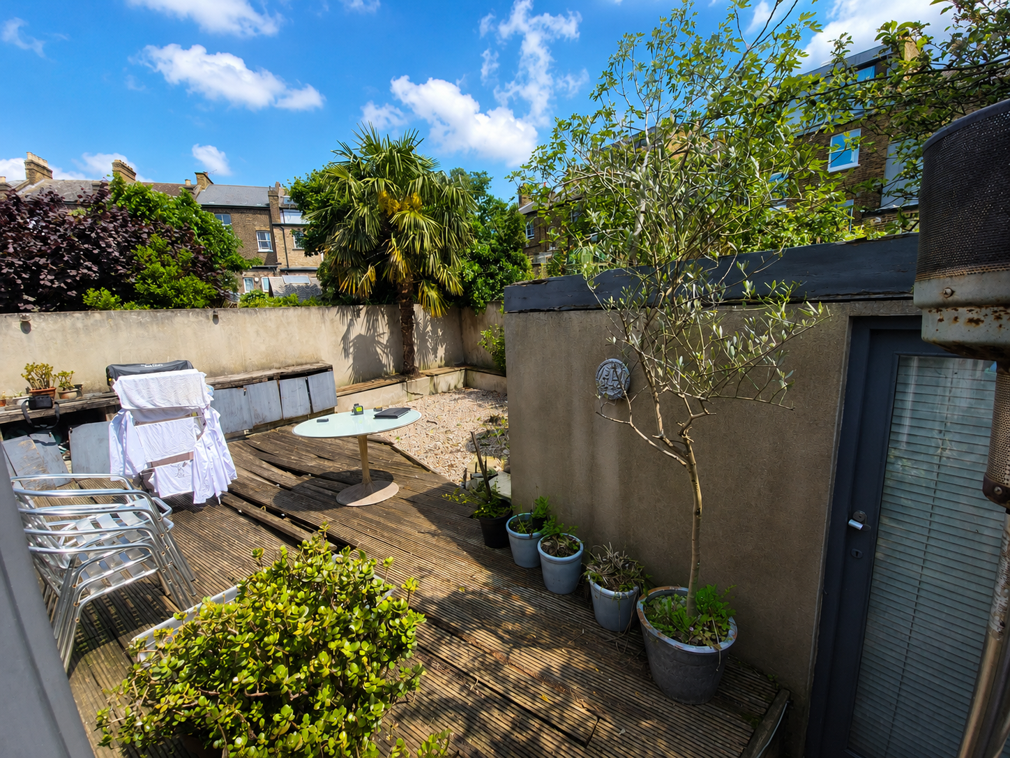 Backyard patio area with wooden deck, potted plants, a small round table, stacked chairs, and trees under a bright blue sky with some clouds.
