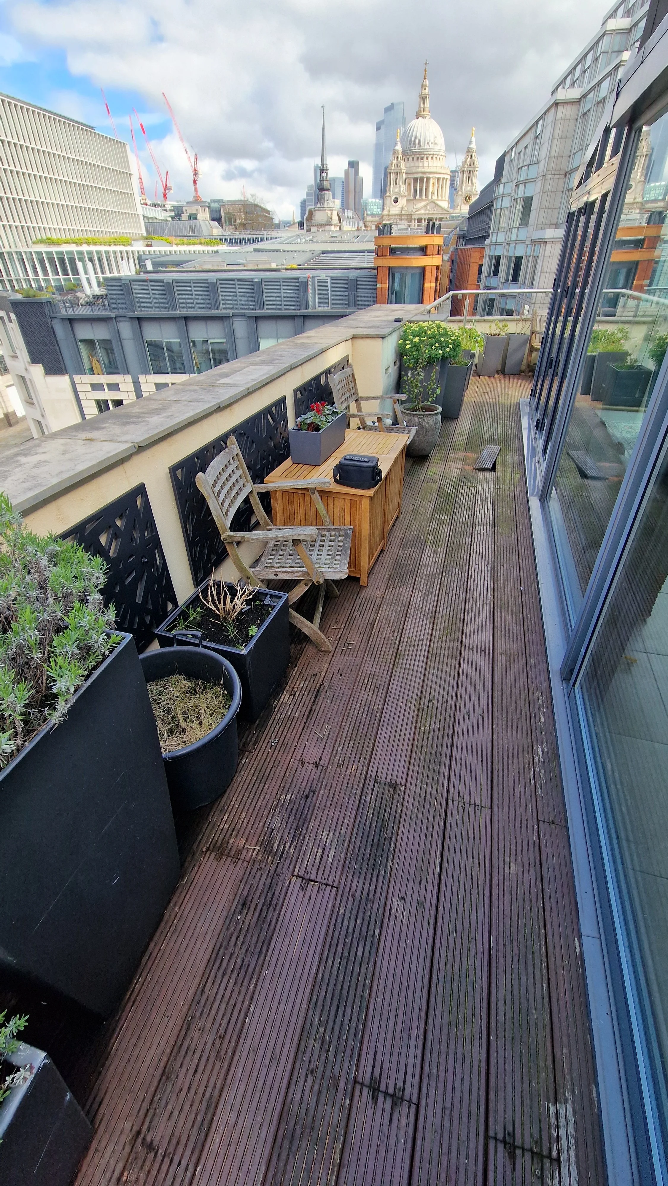 City rooftop terrace with wooden flooring, potted plants, outdoor chairs, a small wooden table, and a view of historic and modern buildings including St. Paul's Cathedral in London.