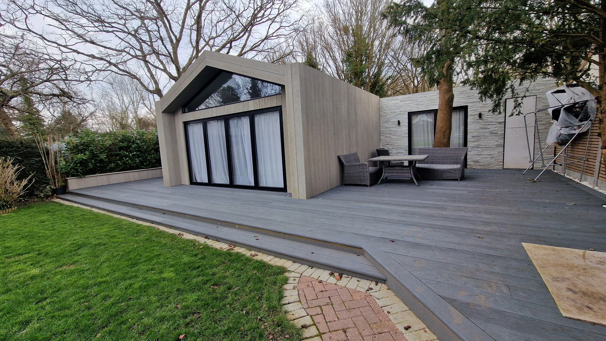 Modern house with large glass sliding doors, outdoor patio furniture, and wooden deck, surrounded by trees and greenery.